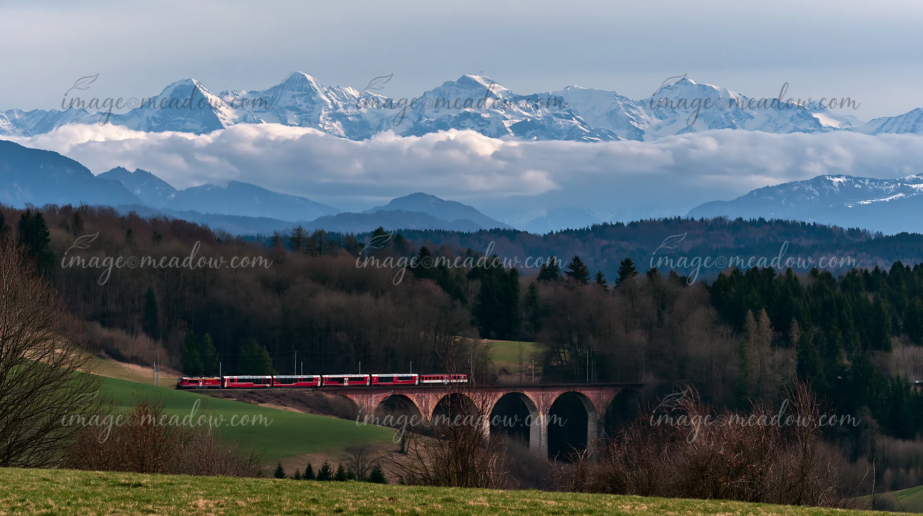 A train crosses a stone viaduct with dramatic clouds around alpine peaks in the background.
