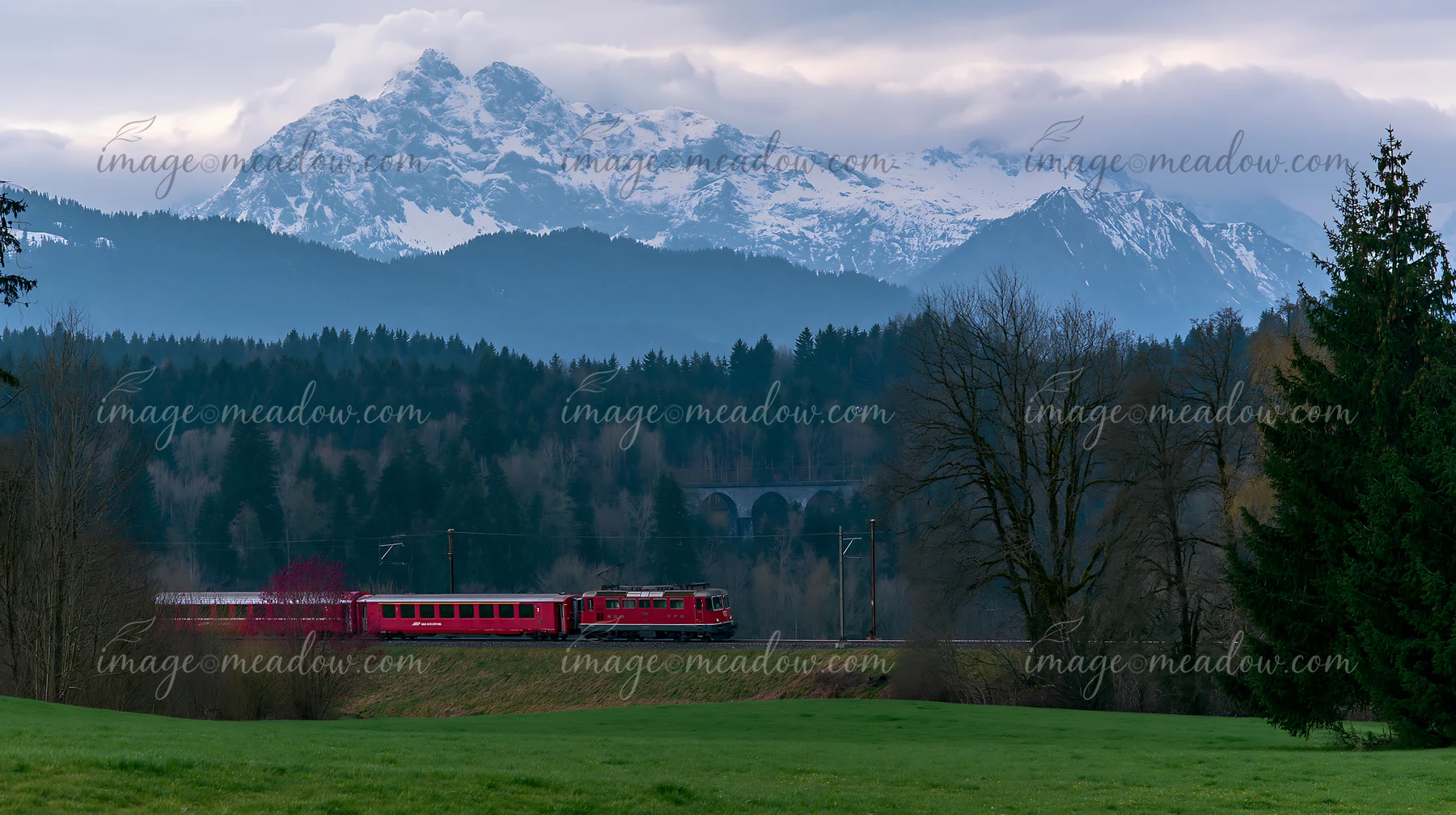 A train travels along a track with dramatic clouds around alpine peaks in the background.