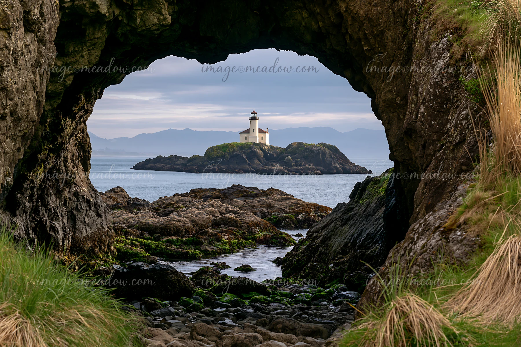 A view of a coastline through a sea cave reveals a lighthouse on a small island.