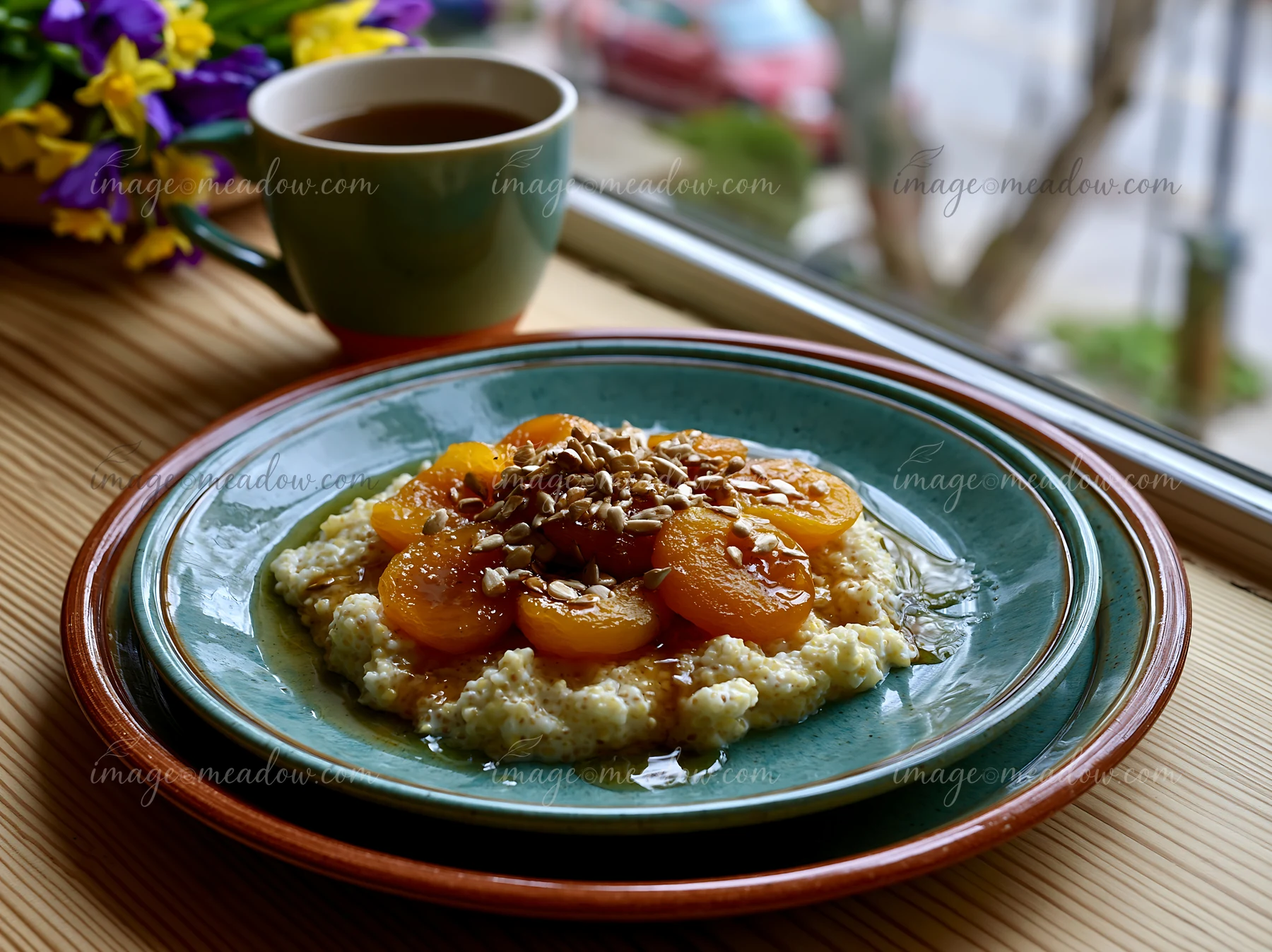 A warm bowl of oats topped with apricots and seeds sits on a table near a window, accompanied by a cup of tea.