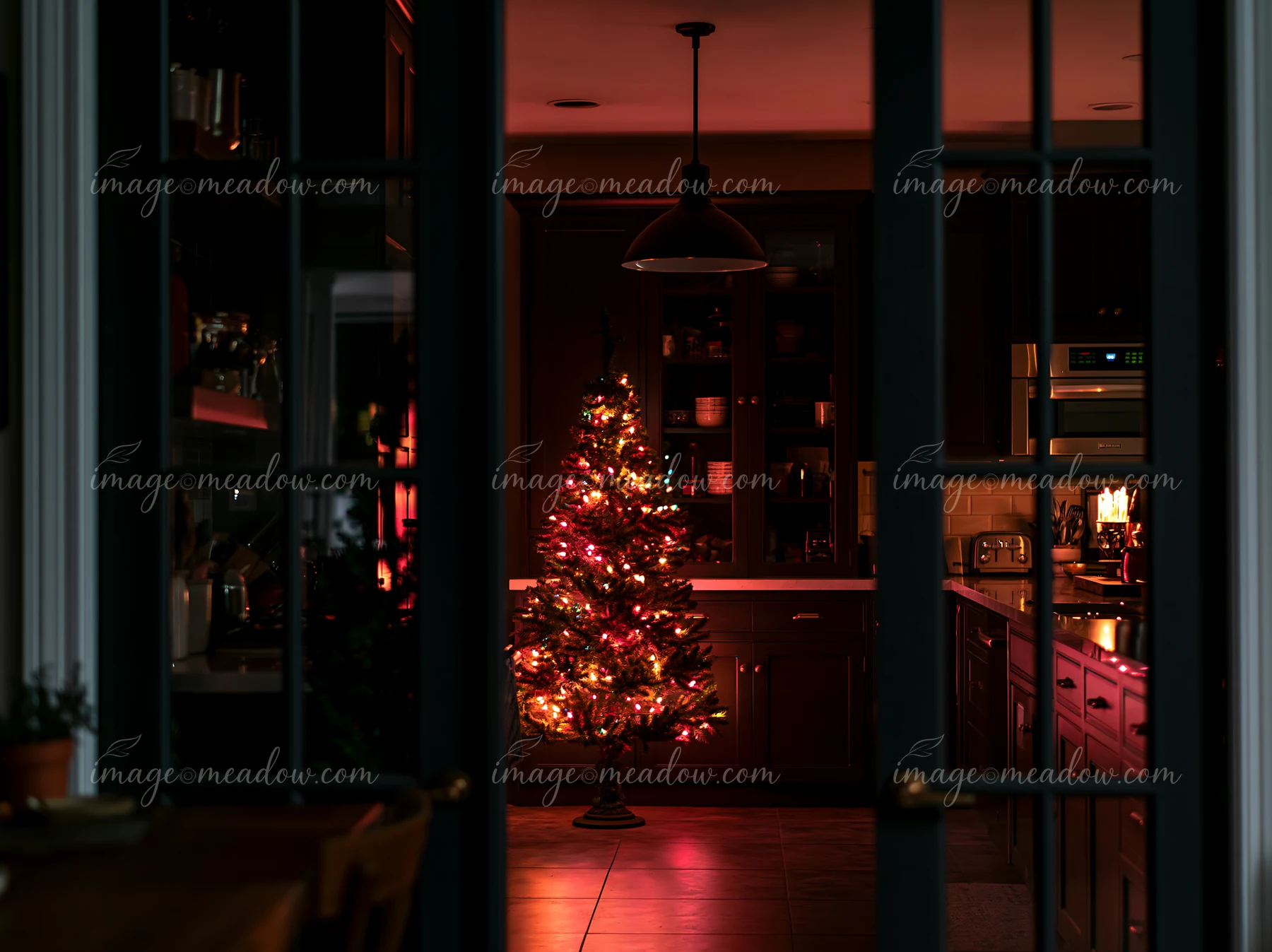 A warm light shines in a modern kitchen where a Christmas tree reflects in the glass doors.