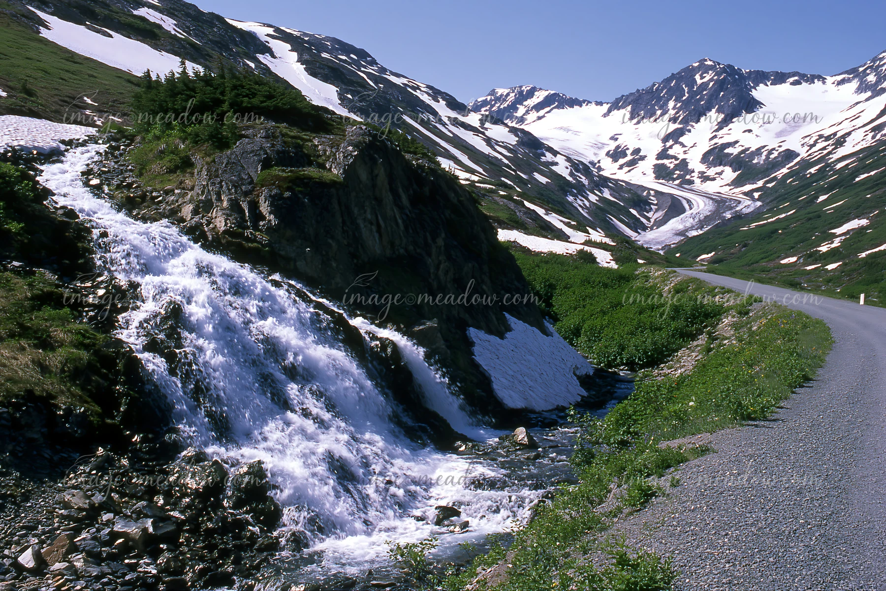A waterfall cascades over rocky terrain with snow-capped mountains in the background.