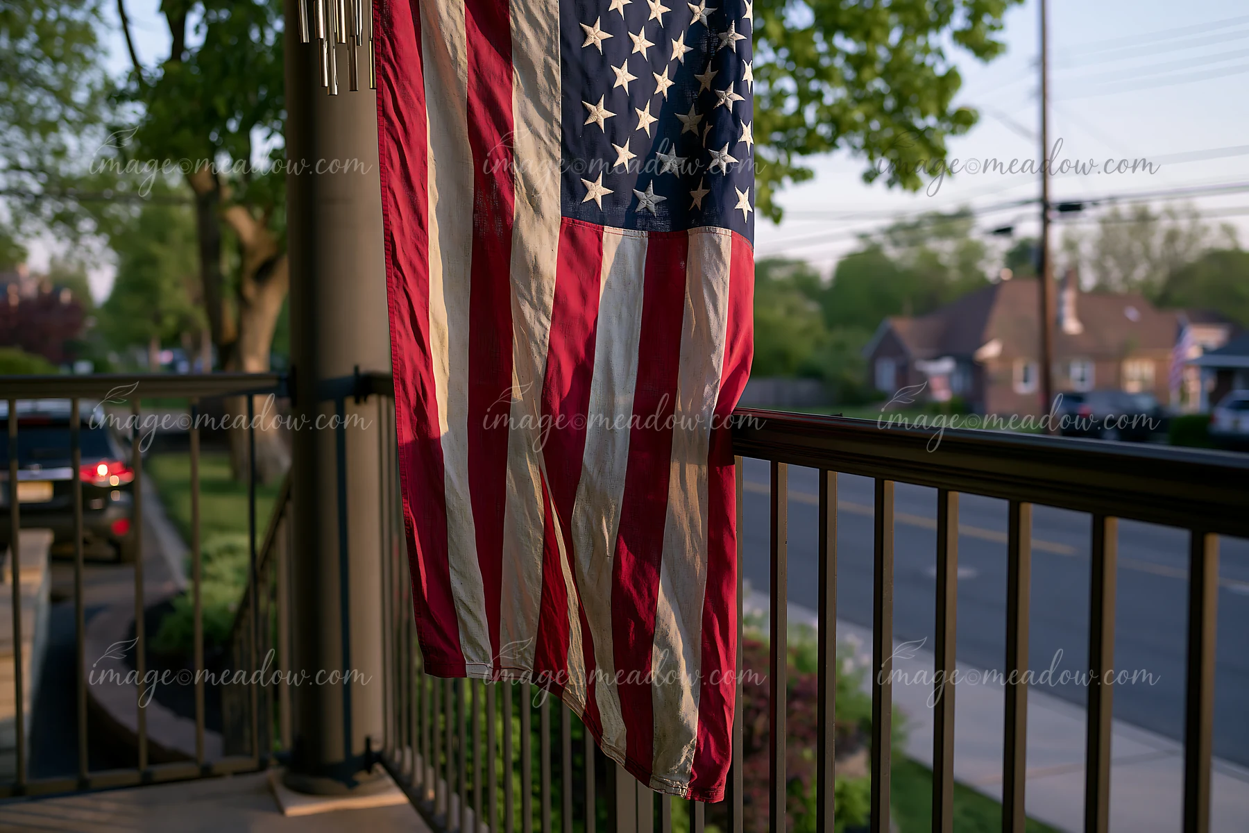 A weathered American flag hangs from a porch railing, waving gently in a suburban neighborhood.