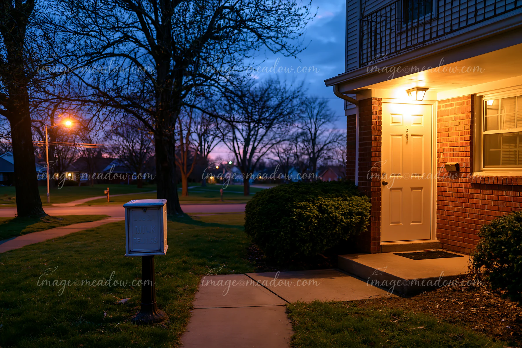 A well-lit apartment doorstep features an insulated milk delivery box, surrounded by grass and shrubbery, during the early evening hours.