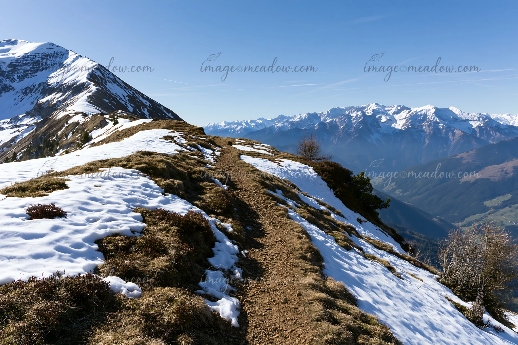 A winding trail stretches along a high mountain ridge with snow patches.