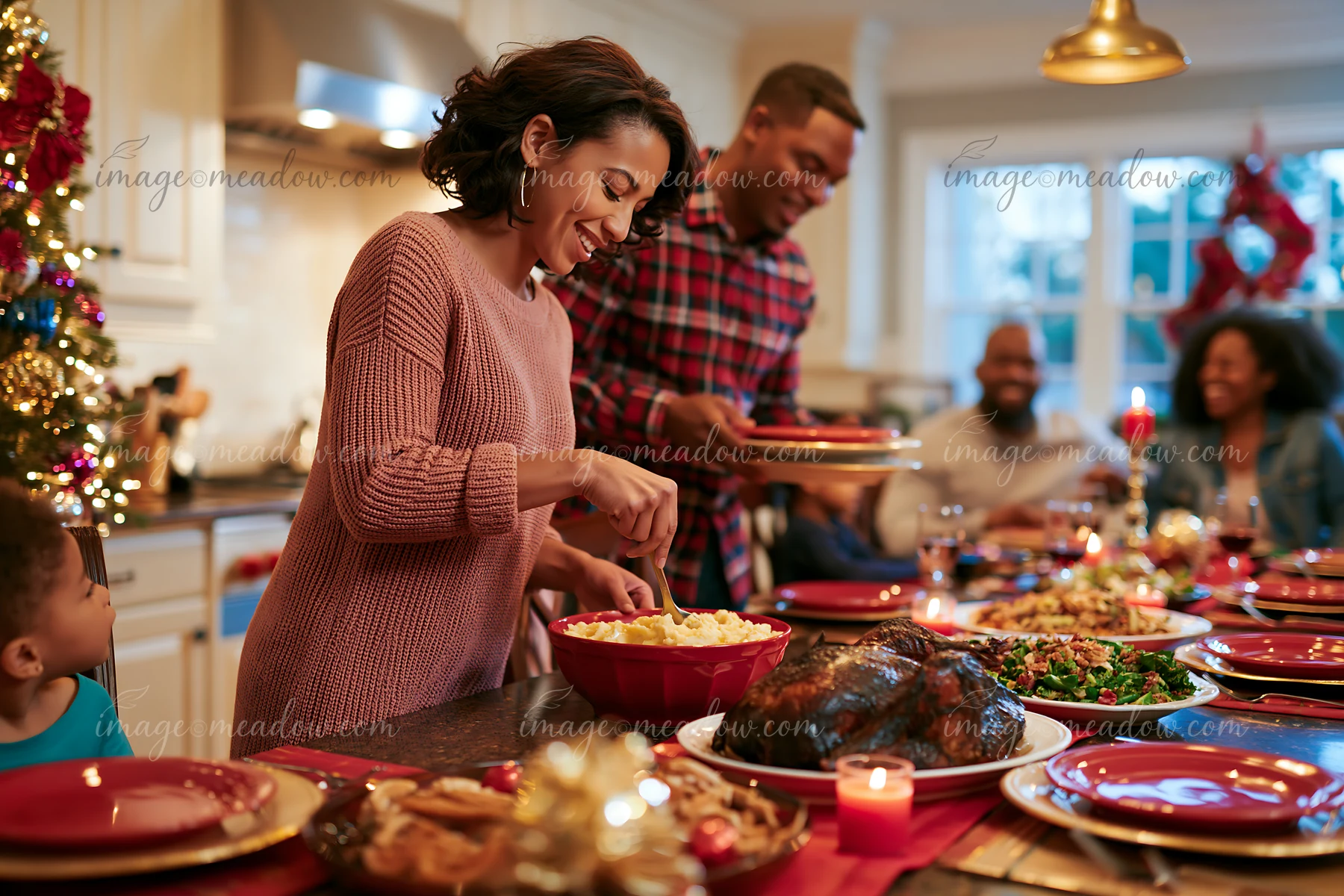 A woman and a man serve food at a Christmas dinner table while family members sit nearby.