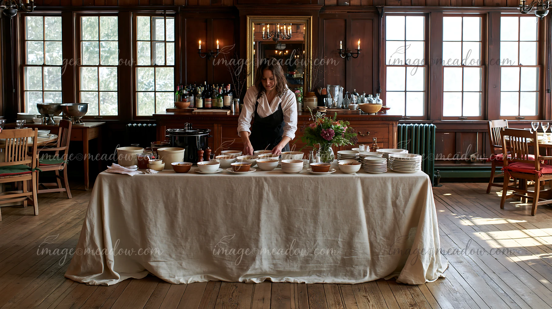 A woman arranges bowls on a rustic wooden buffet covered with a linen tablecloth in a dining room.