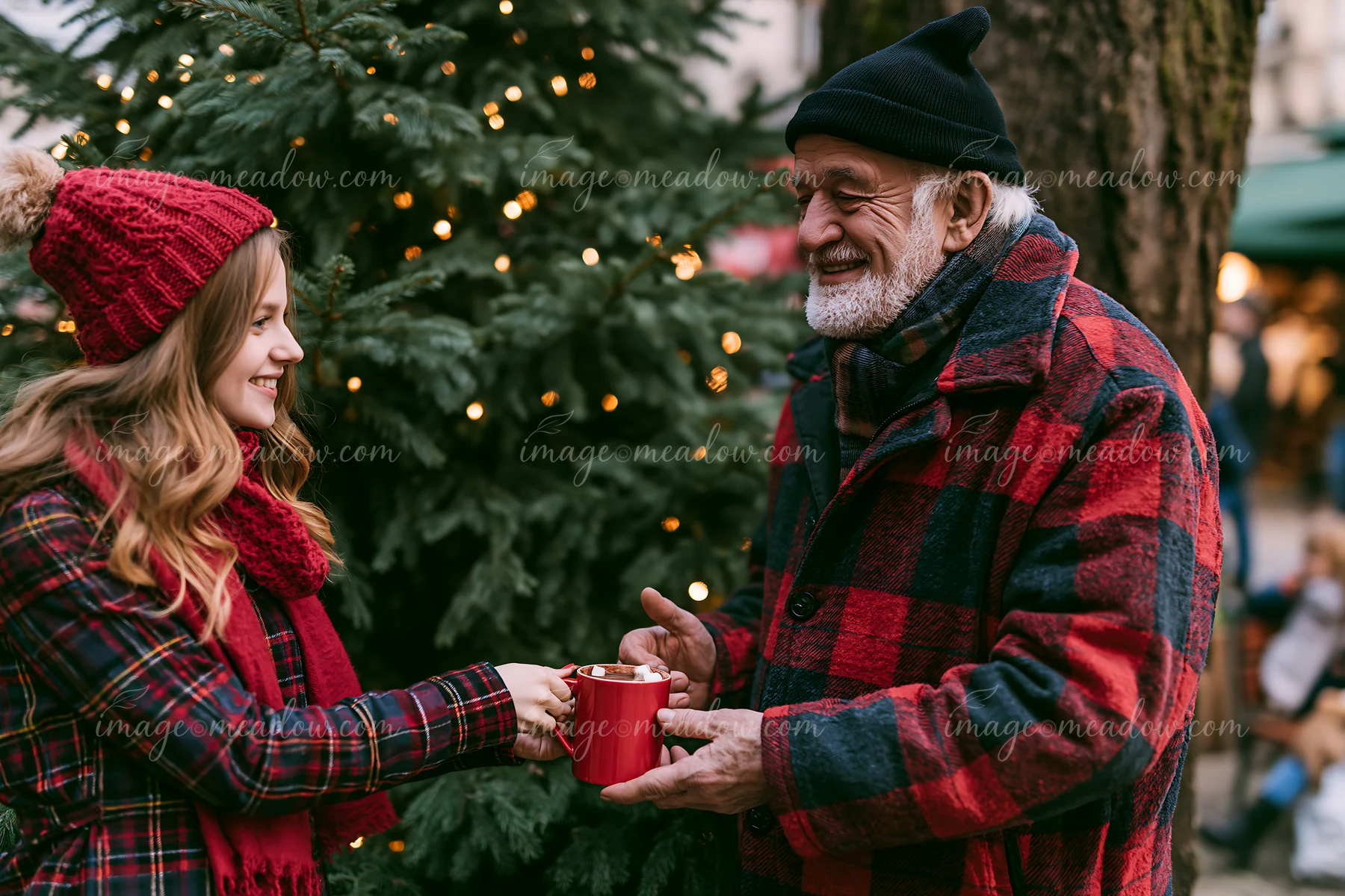 A woman hands a red mug of warm cocoa topped with marshmallows to an elderly man during a festive gathering near a decorated tree.