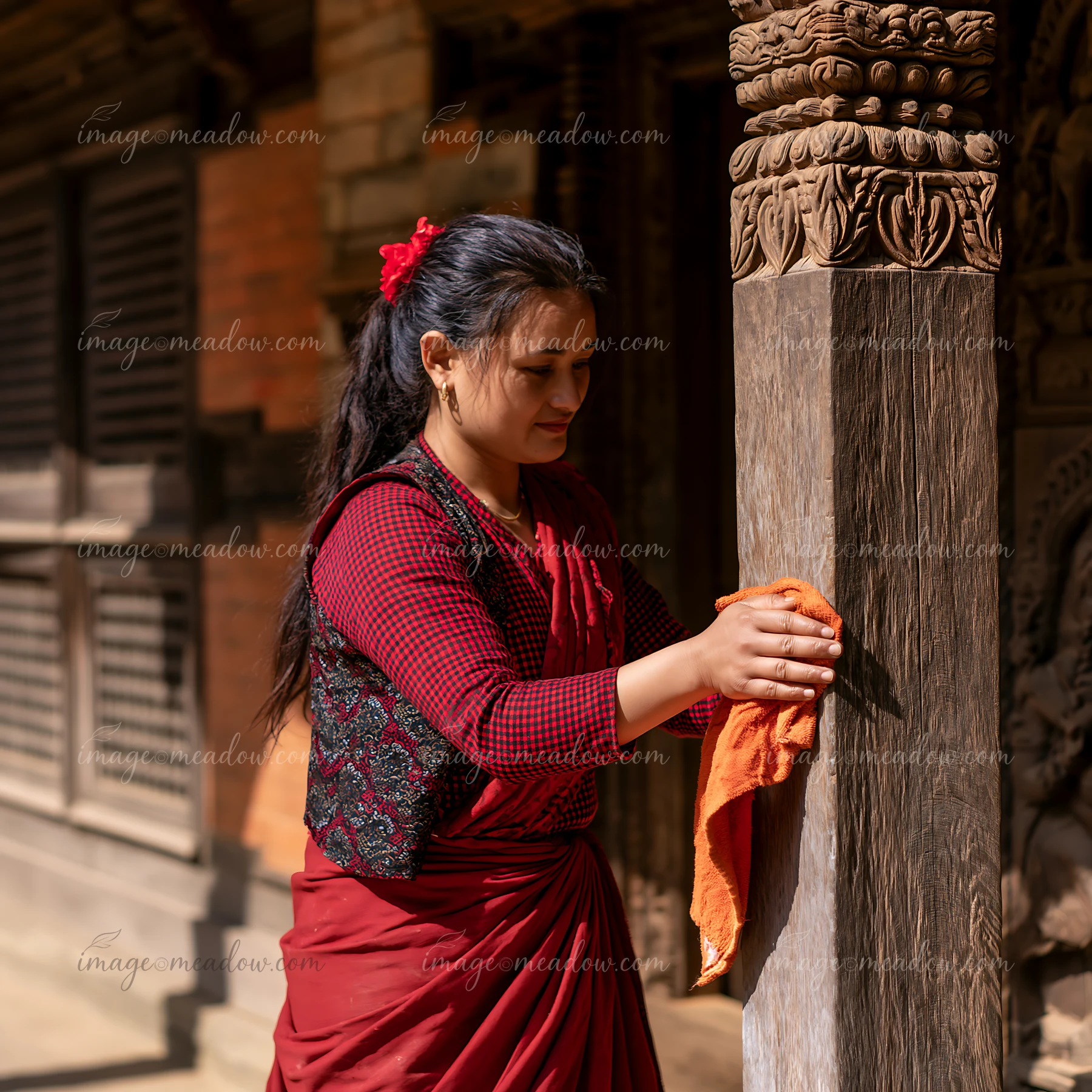 A woman in heritage attire is cleaning a wooden post of a traditional building in a sunny outdoor setting.
