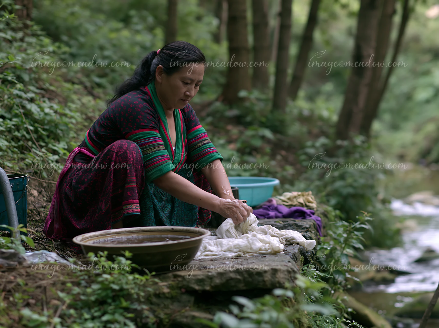 A woman in traditional clothing washes linens by a stream surrounded by trees.