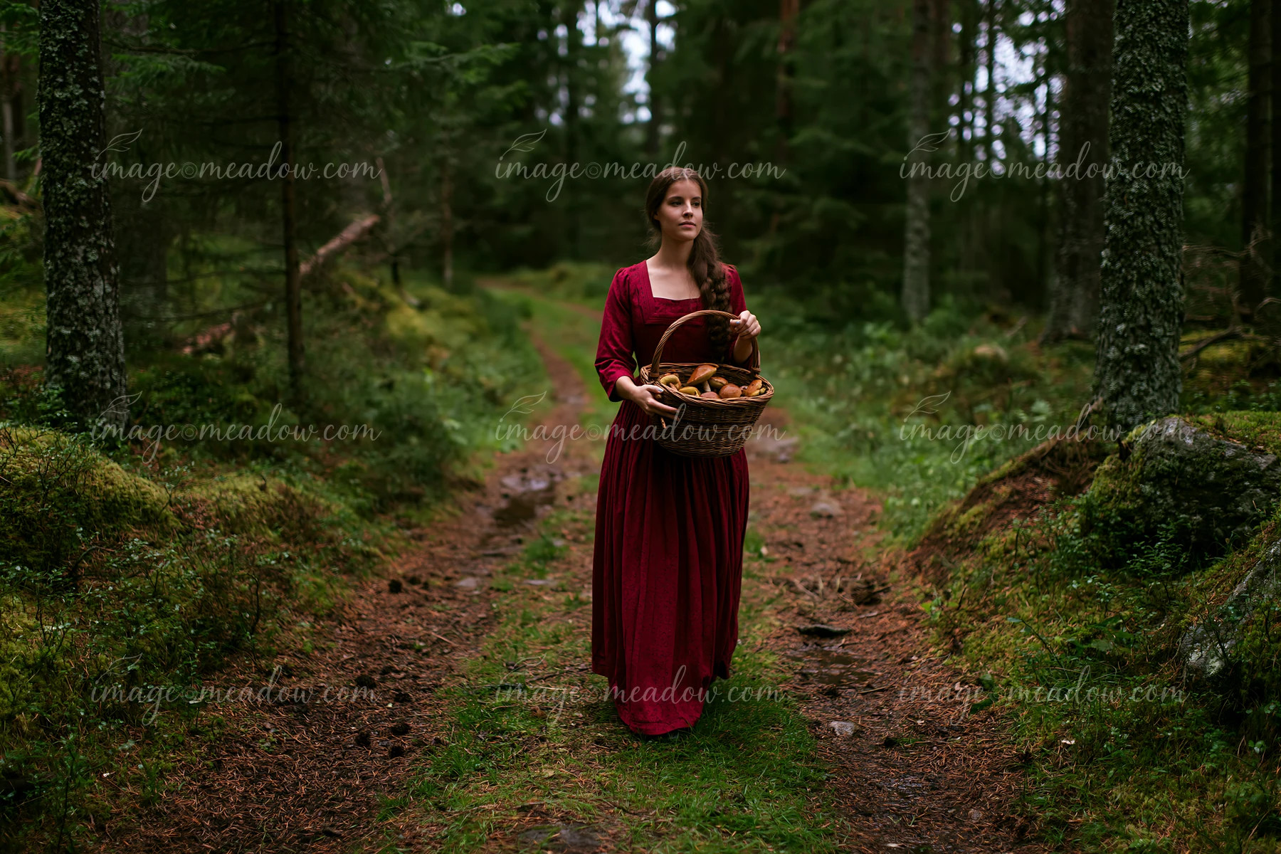A woman in traditional dress walks along a forest path, carrying a basket of mushrooms.
