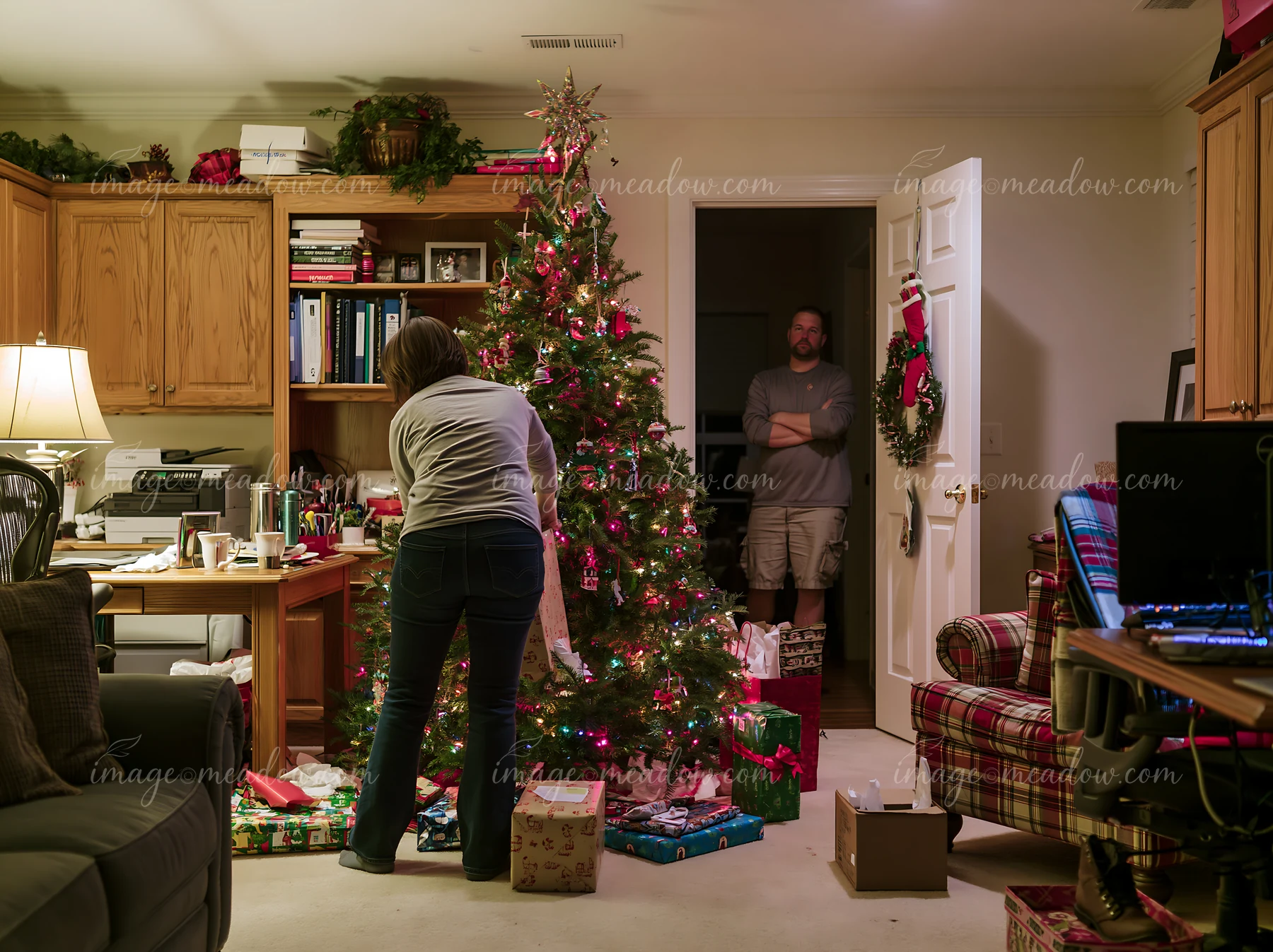 A woman is arranging gifts around a Christmas tree in a home office.
