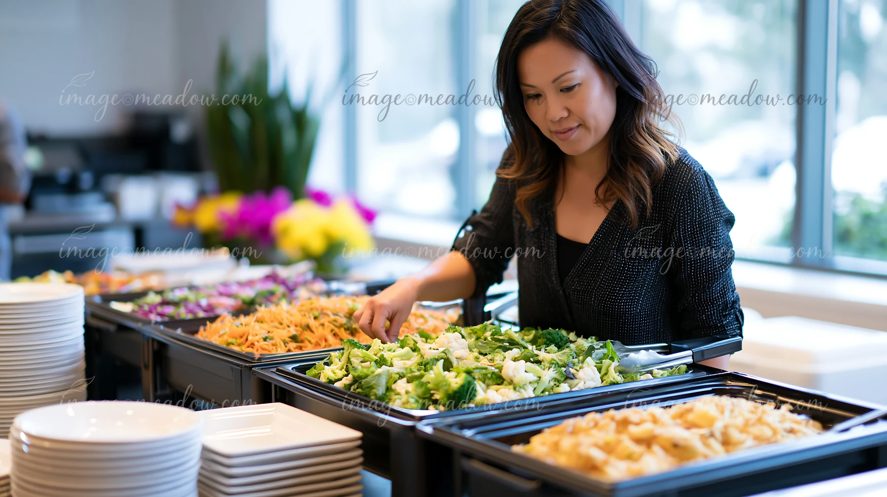 A woman is choosing fresh salad ingredients from a corporate lunch buffet in a bright setting.