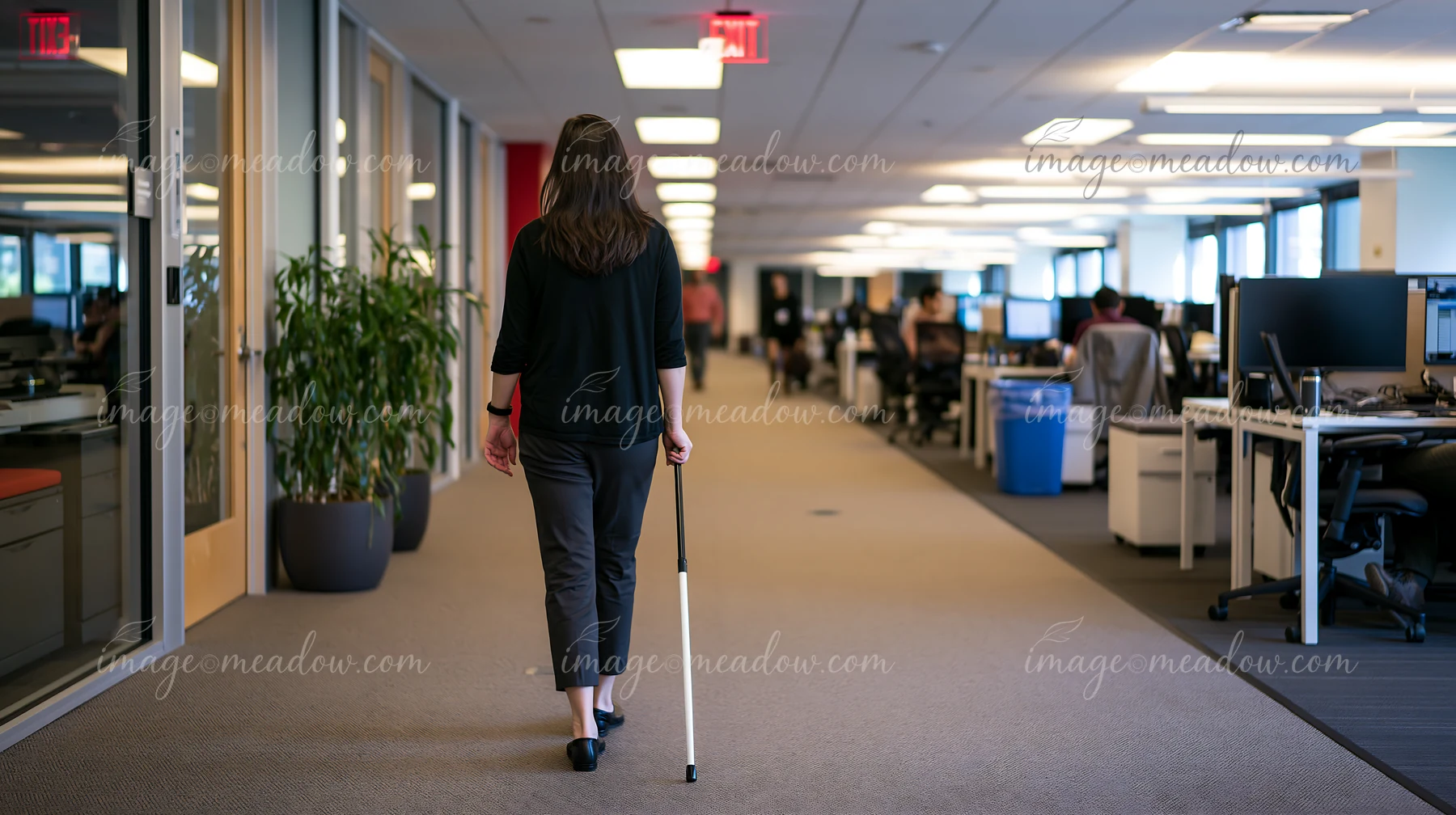 A woman uses a white cane to navigate through an office environment with cubicles and computer workstations.