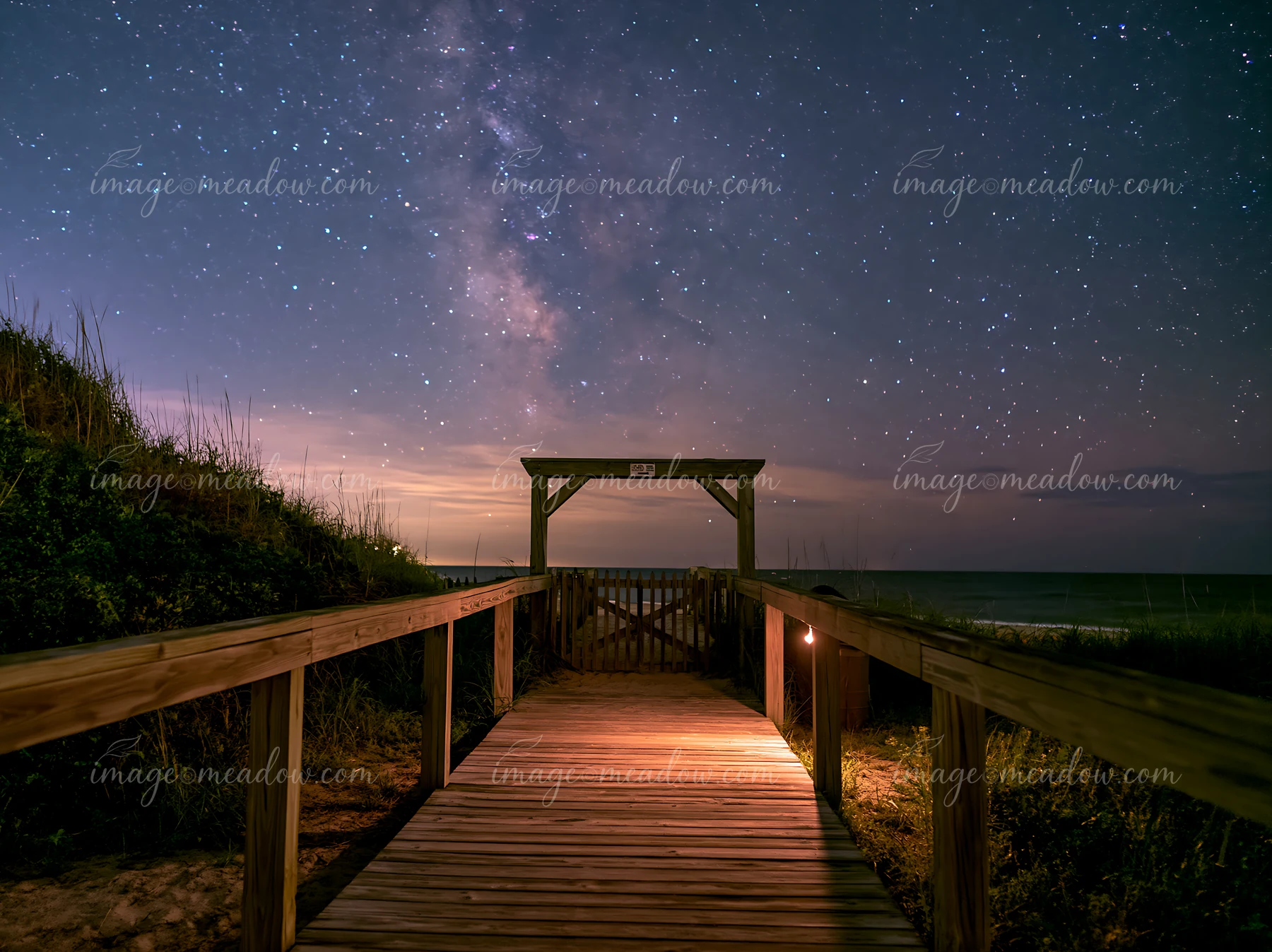 A wooden boardwalk leads to the beach under a night sky filled with stars.