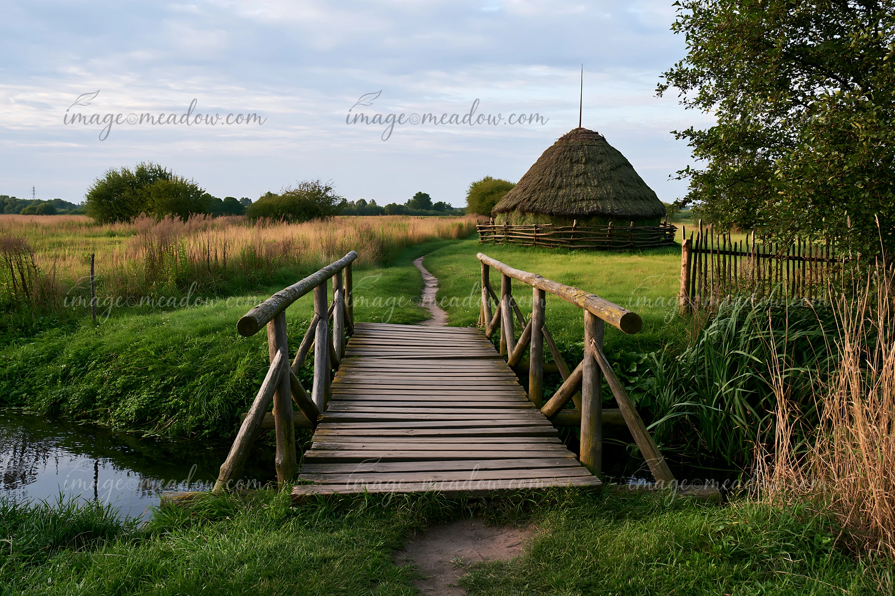 A wooden bridge crosses a small stream in a green meadow with a traditional haystack in the background.