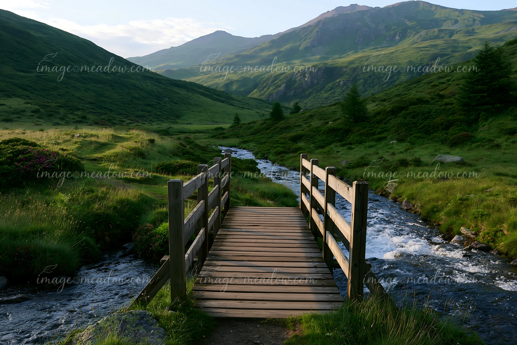 A wooden bridge spans a stream, surrounded by hills and greenery under golden light.