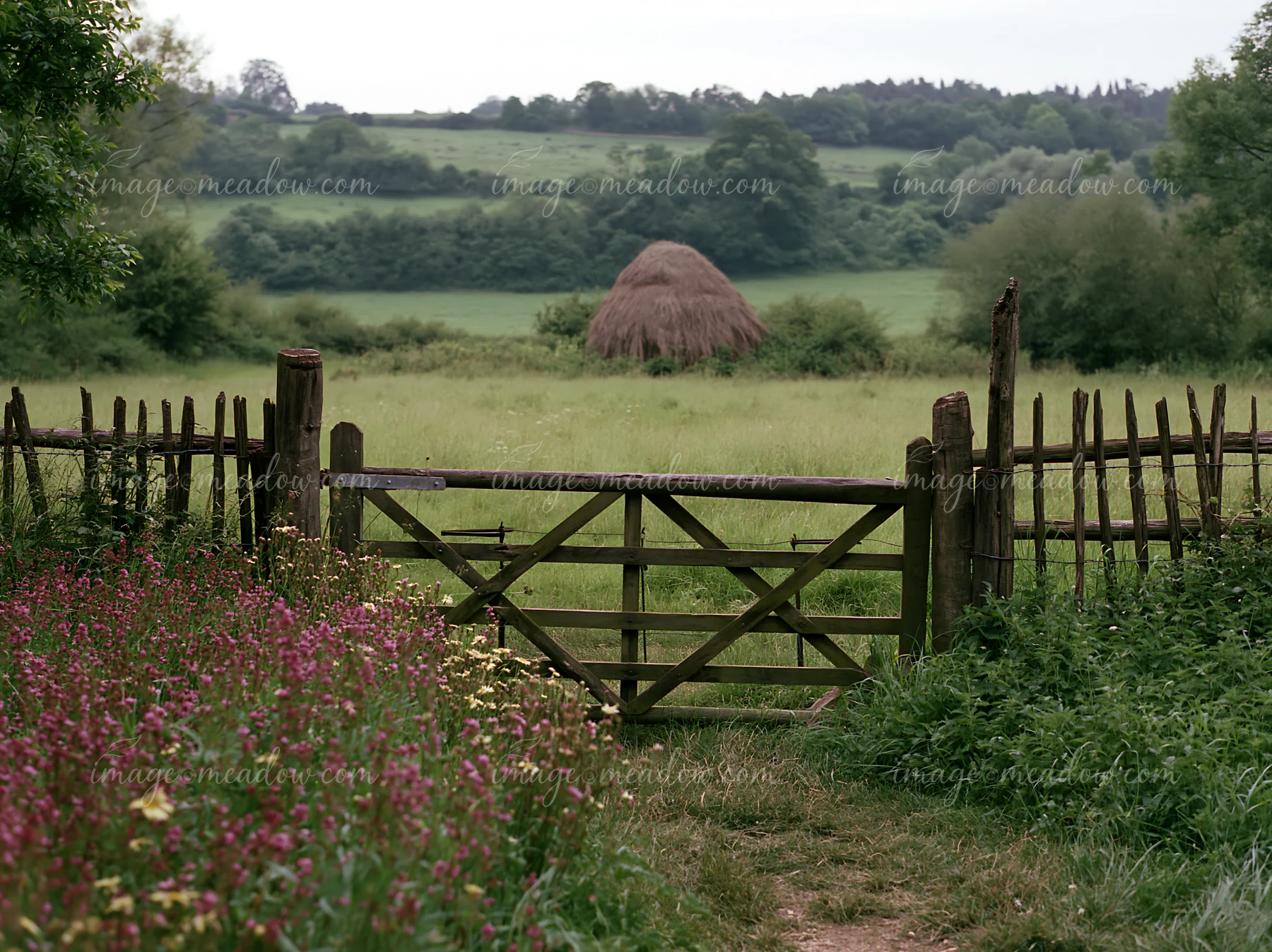 A wooden gate opens to a green meadow with a traditional haystack in the background.