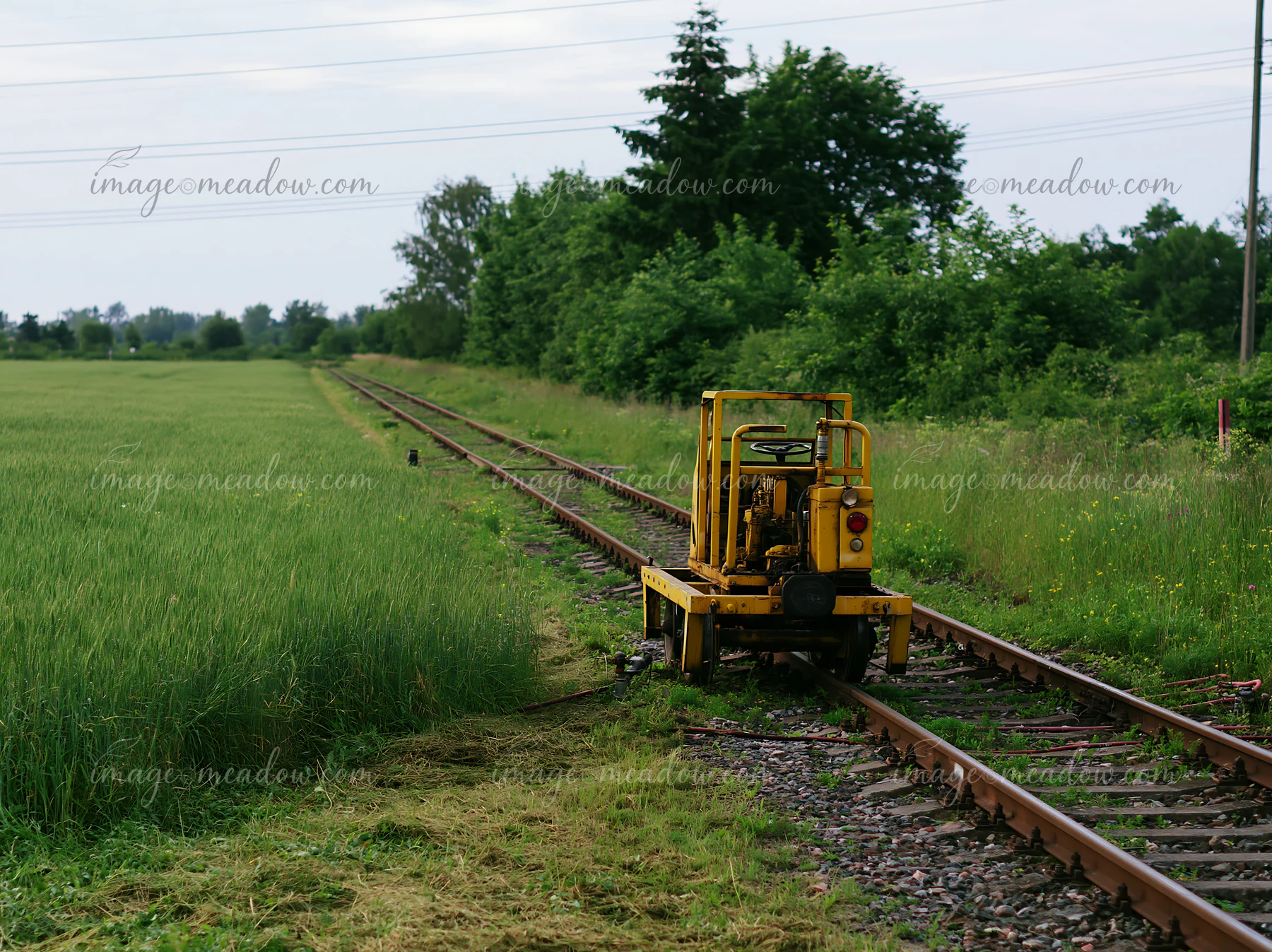 A yellow maintenance vehicle is working on the railway tracks next to a wide meadow alongside railway tracks.