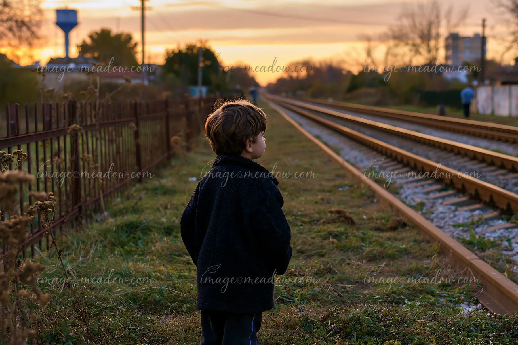 A young boy stands in a meadow alongside railway tracks, looking towards the horizon.