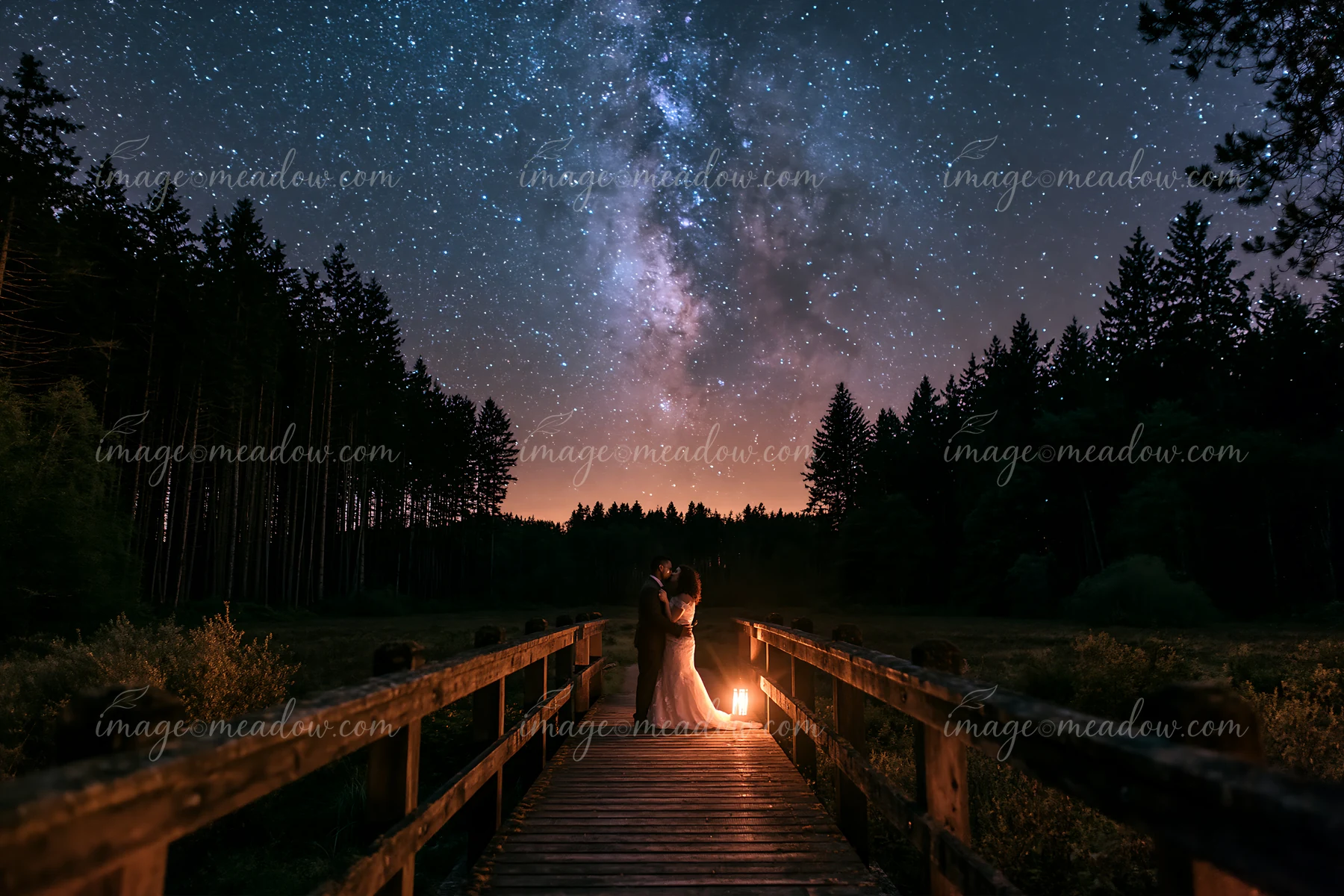 A young couple kissing beneath visible Milky Way on a wooden bridge surrounded by trees.