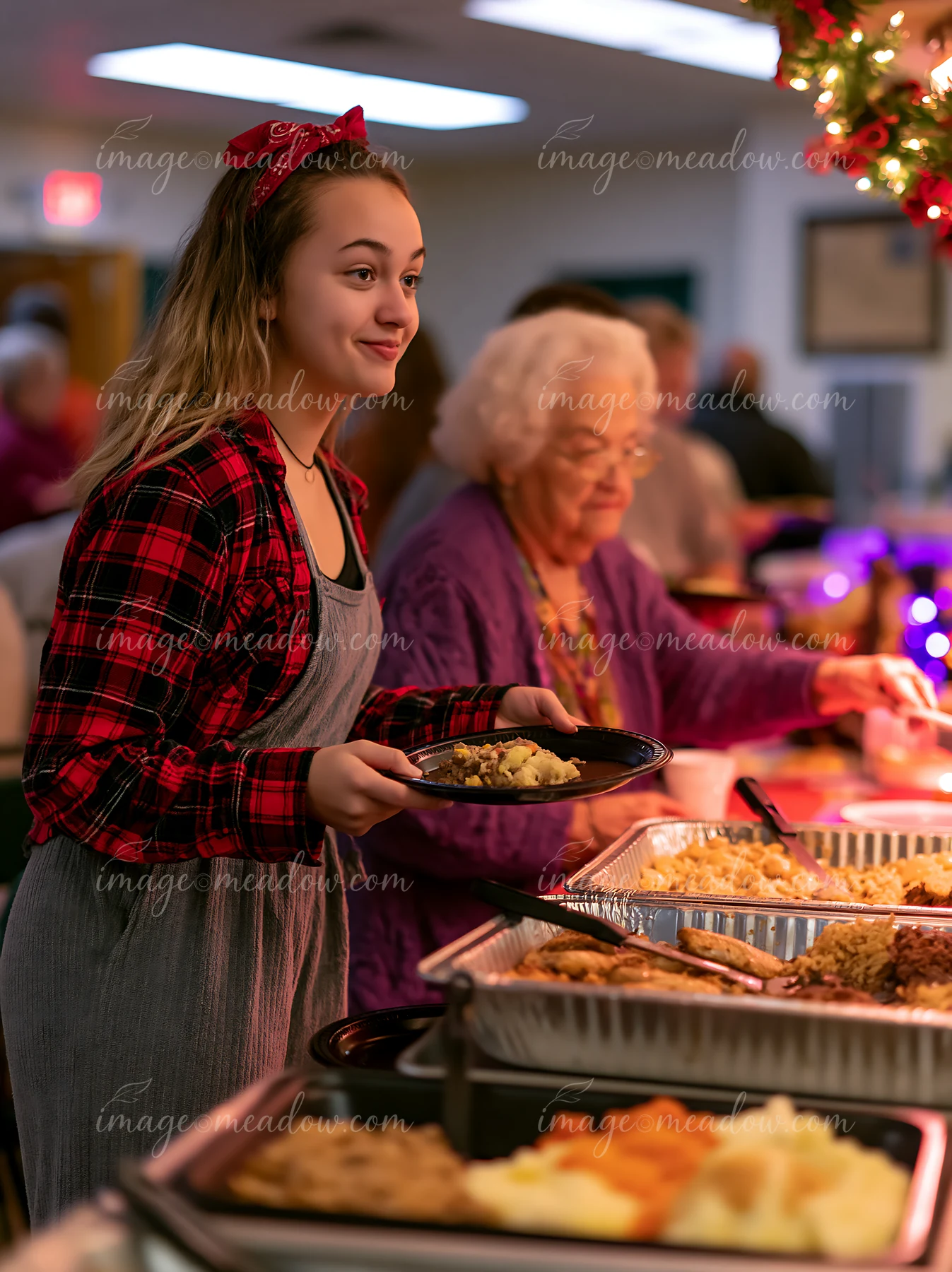 A young woman holds a plate as she serves food at a community potluck buffet in a warm setting, surrounded by people enjoying the meal and socializing.