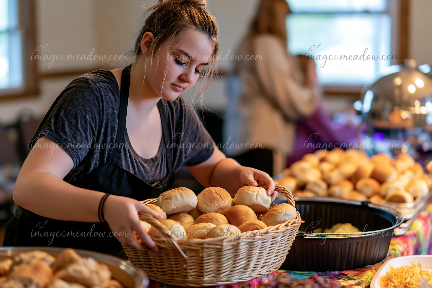 A young woman places freshly baked rolls into a basket while assisting at a charity event buffet.