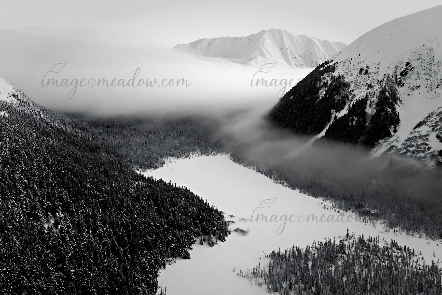 An aerial view of a snowy meadow valley, surrounded by mountains, with mist hovering above the landscape, adding depth and dimension to the view. Presented as a