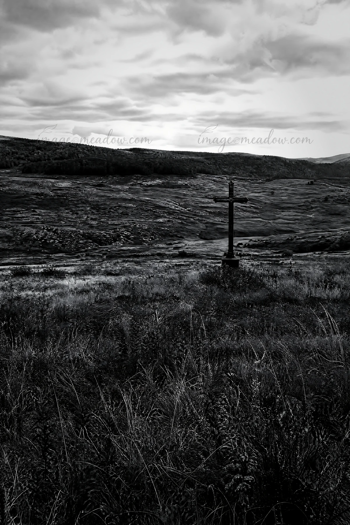 An aged Christian cross stands in a meadow located in a deep mountain valley during autumn. The landscape features hills and patches of grass under a cloudy sky