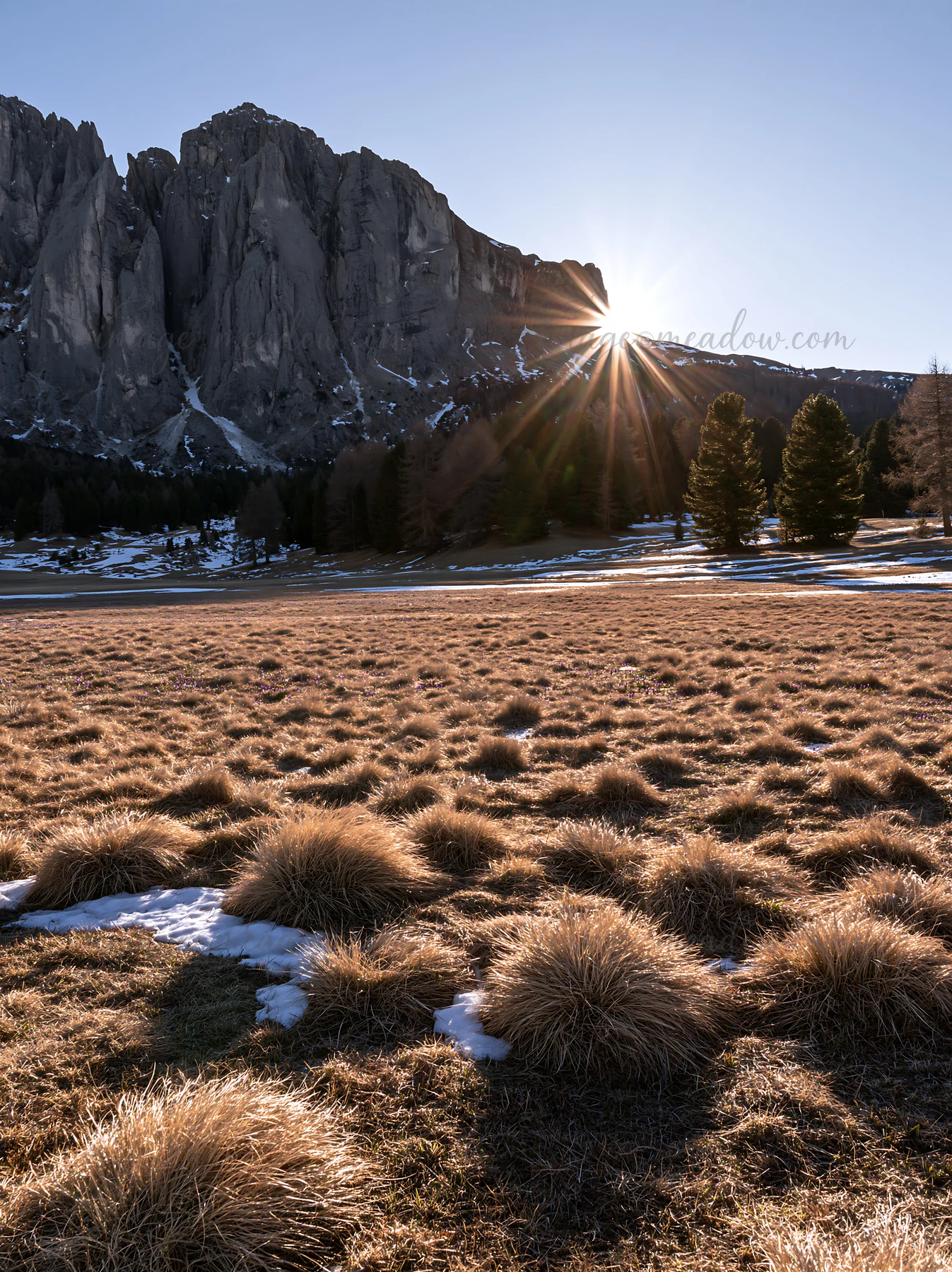 Sunlight breaks over the Alpe di Siusi meadow, illuminating the grass and creating shadows on the ground. The rugged mountains provide a dramatic backdrop to th