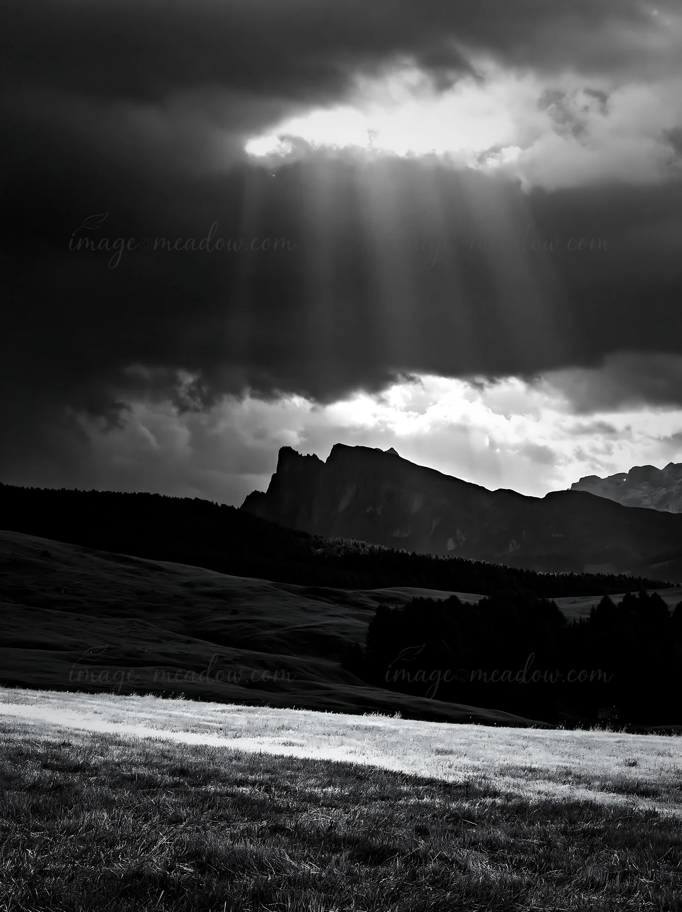 Sunbeams break through dark clouds over the Alpe di Siusi meadow, illuminating the green landscape and nearby mountains. The scene captures a moment of changing
