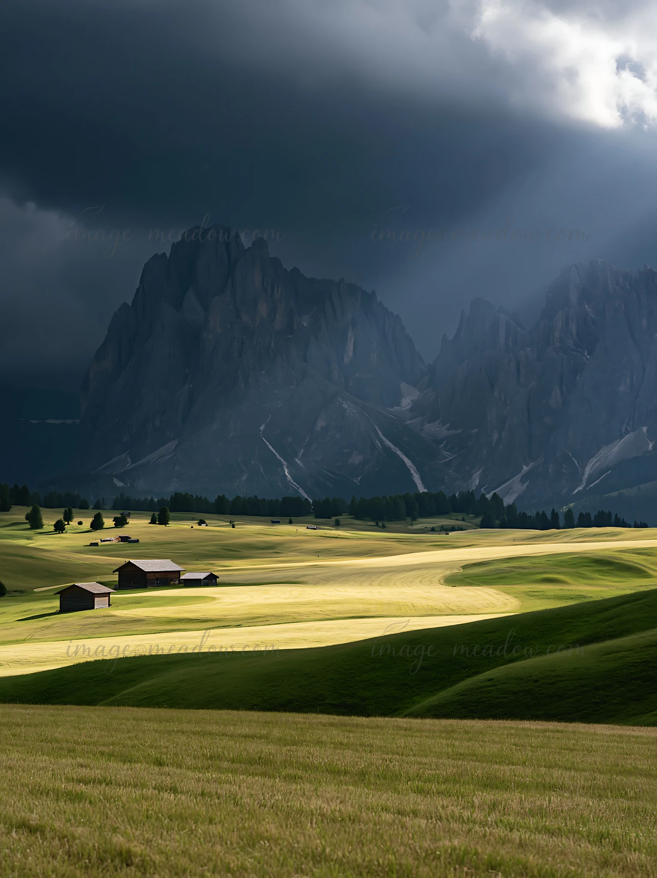 Sunbeams shine down on the Alpe di Siusi meadow. The scene features green grass and wooden structures against a dramatic mountain backdrop.