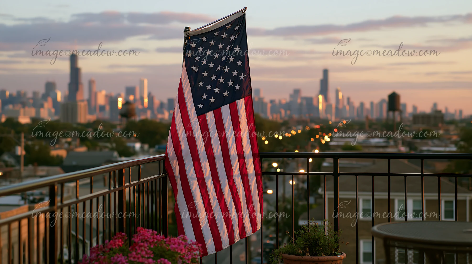 American flag hanging vertically on a balcony with a city skyline in the background during sunset.