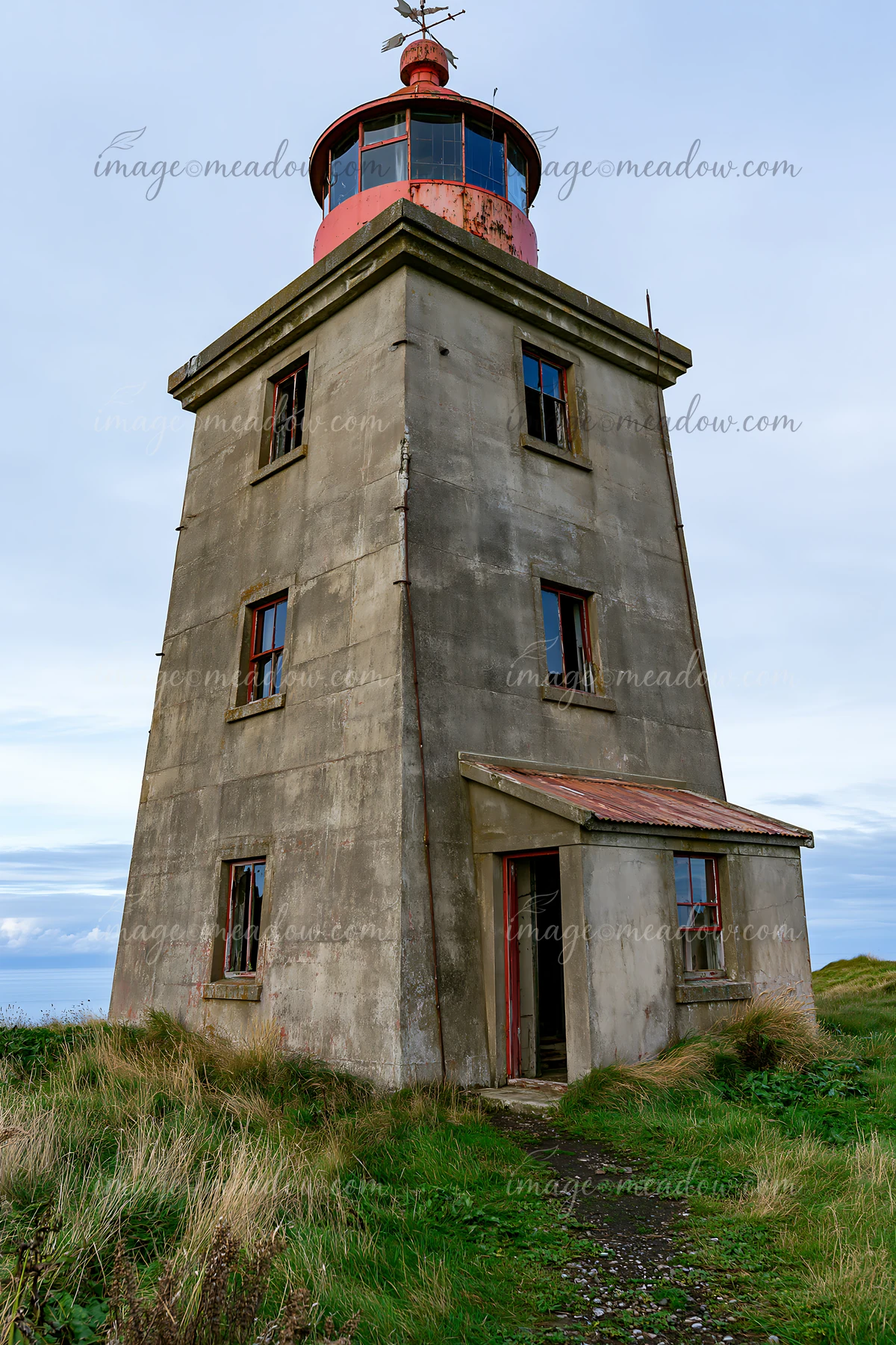 An abandoned lighthouse on a remote coast stands tall and weathered, surrounded by grass and a cloudy sky.