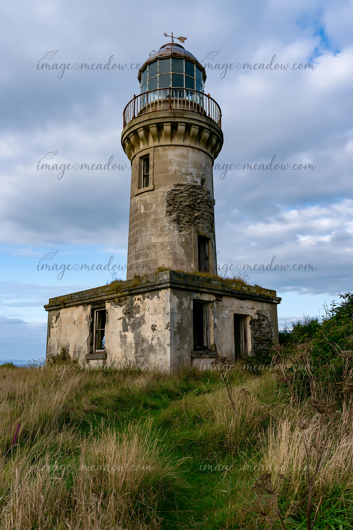 An abandoned lighthouse on a remote coast stands tall with patches of grass surrounding its base.