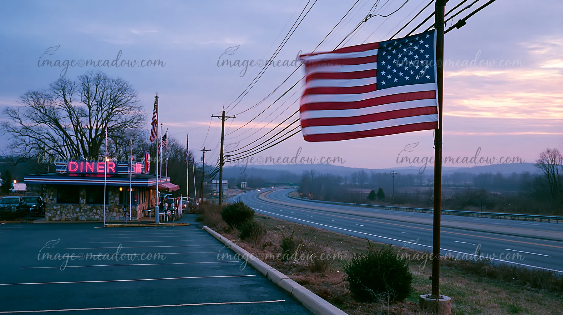 An American flag flying in strong wind stands prominently outside a diner along a quiet highway at dusk.
