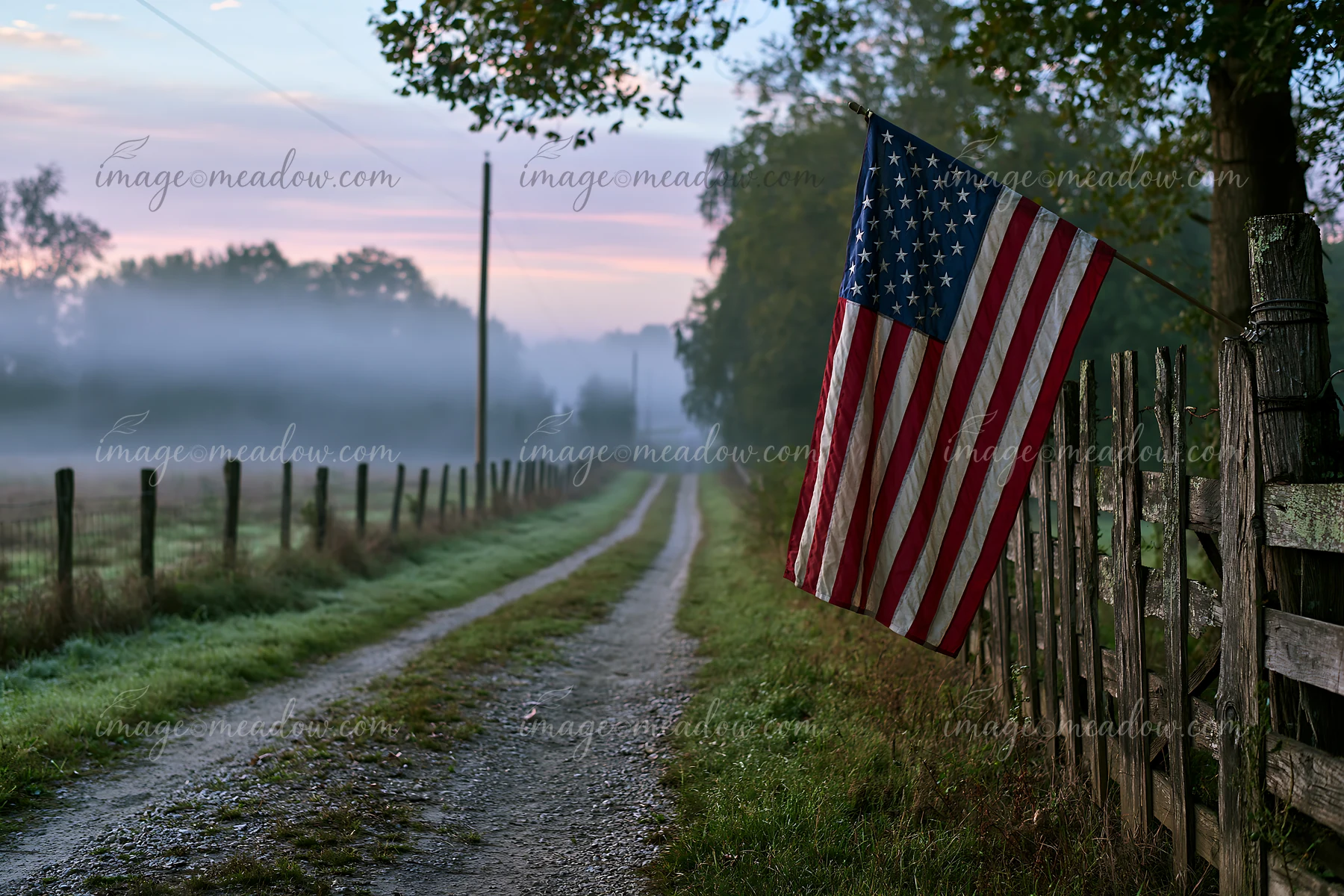 An American flag hangs on a fence by a gravel road surrounded by a misty landscape.