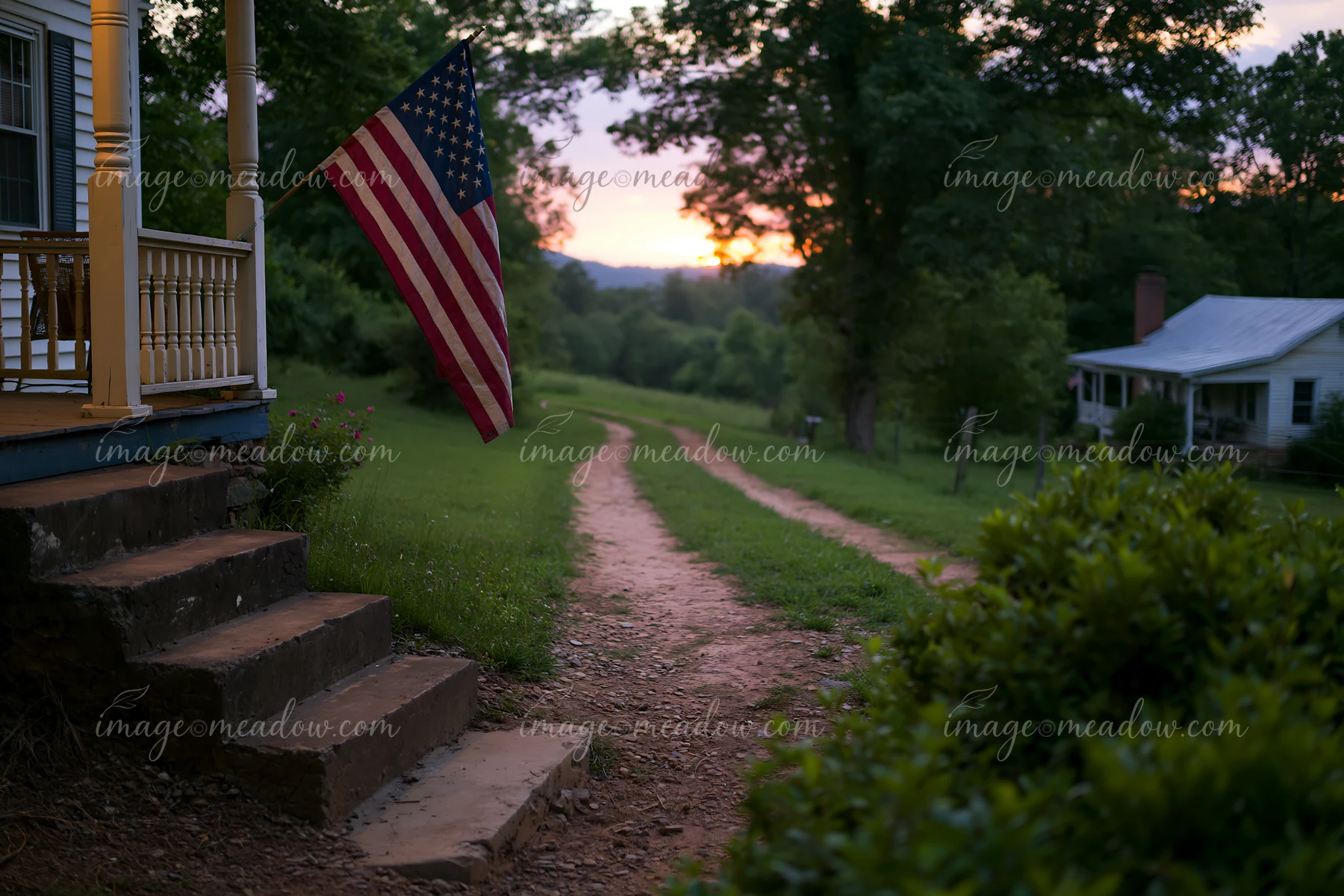 An American flag is displayed on a porch near a rural road as the sun sets behind green trees.