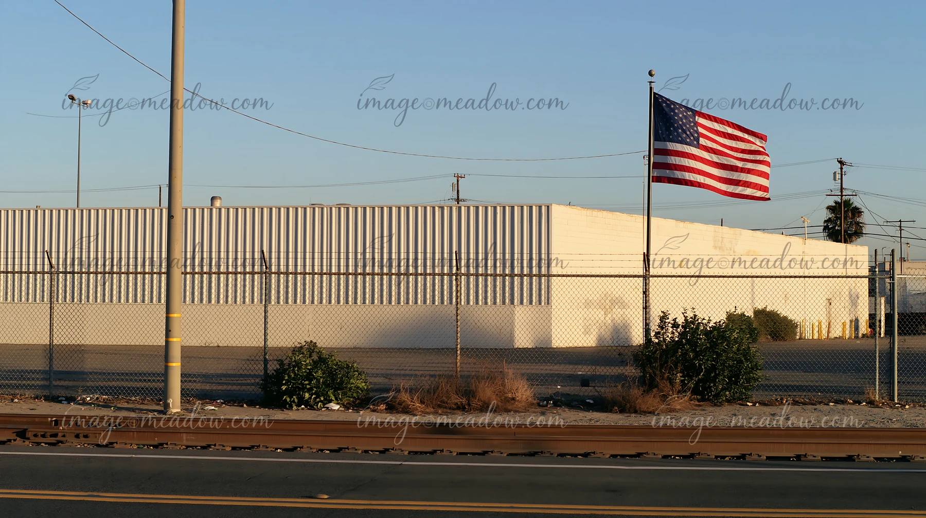 An American flag outside a factory is seen near a fence and road.