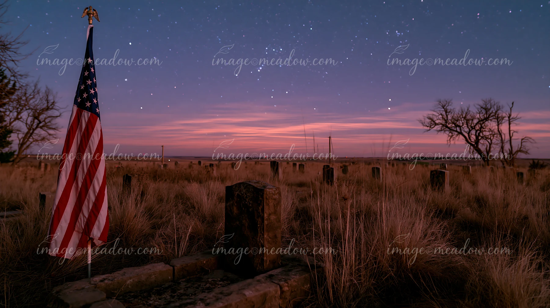 An American flag stands next to grave markers in an open field as stars fill the sky during dusk.