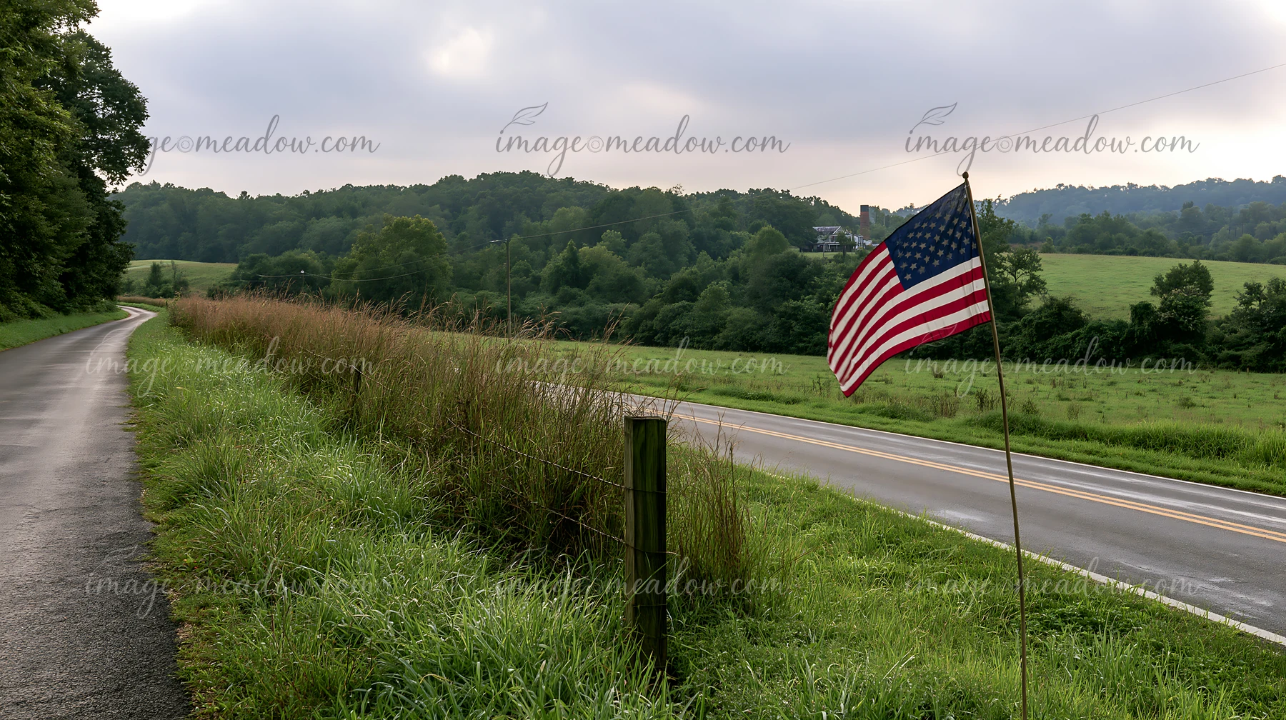An American flag stands tall on a rural road surrounded by green fields and distant hills.