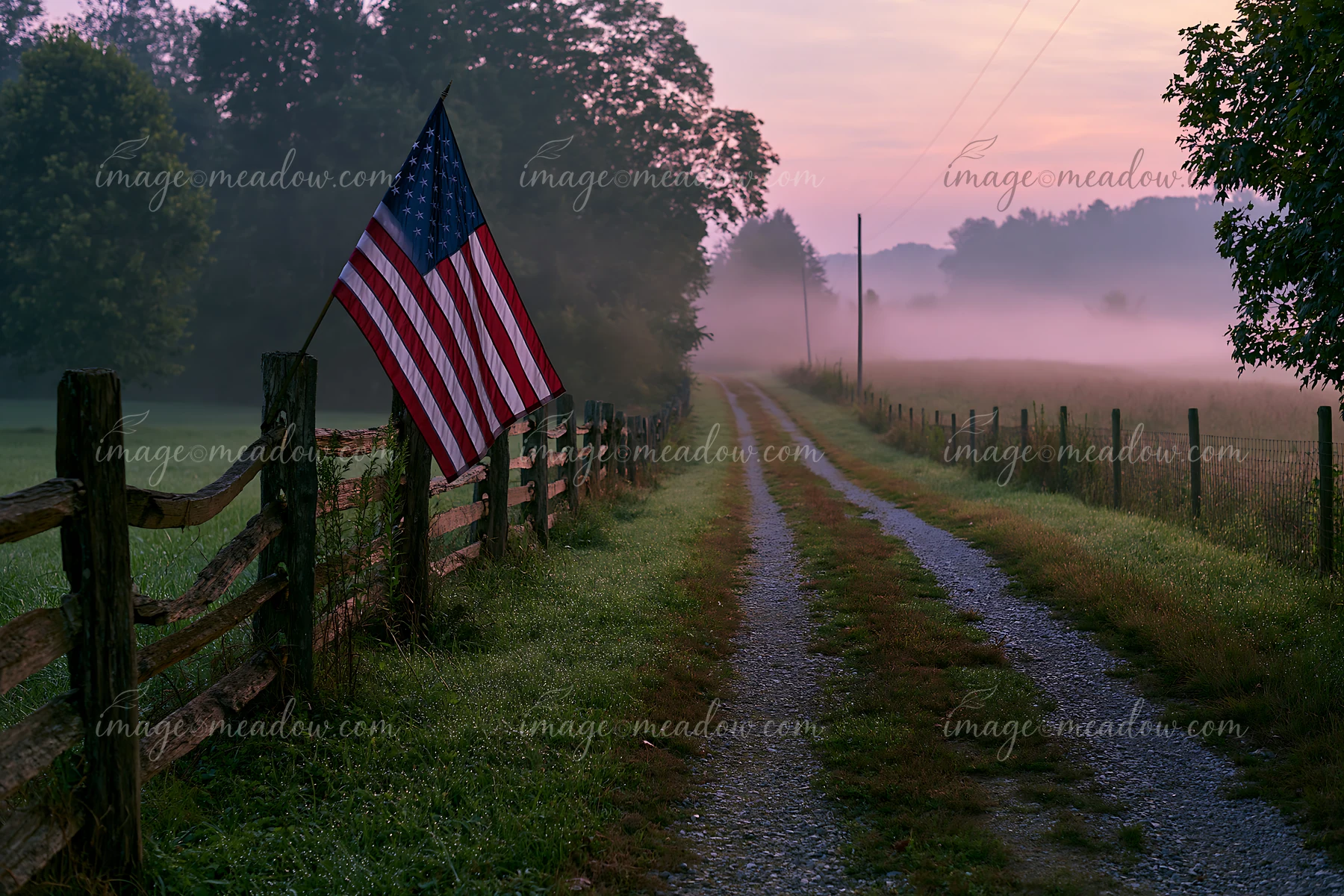 An American flag waves gently beside a gravel road lined with a wooden fence.