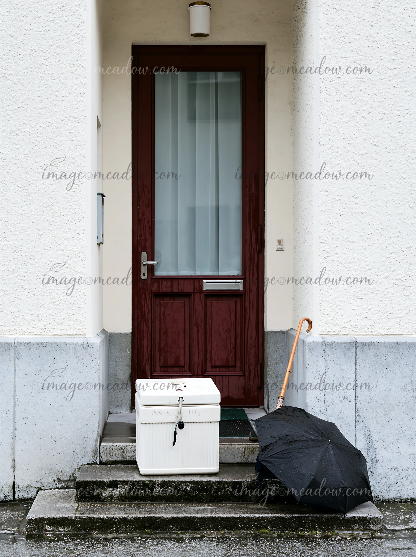 An apartment doorstep shows an insulated milk delivery box placed next to a black umbrella.