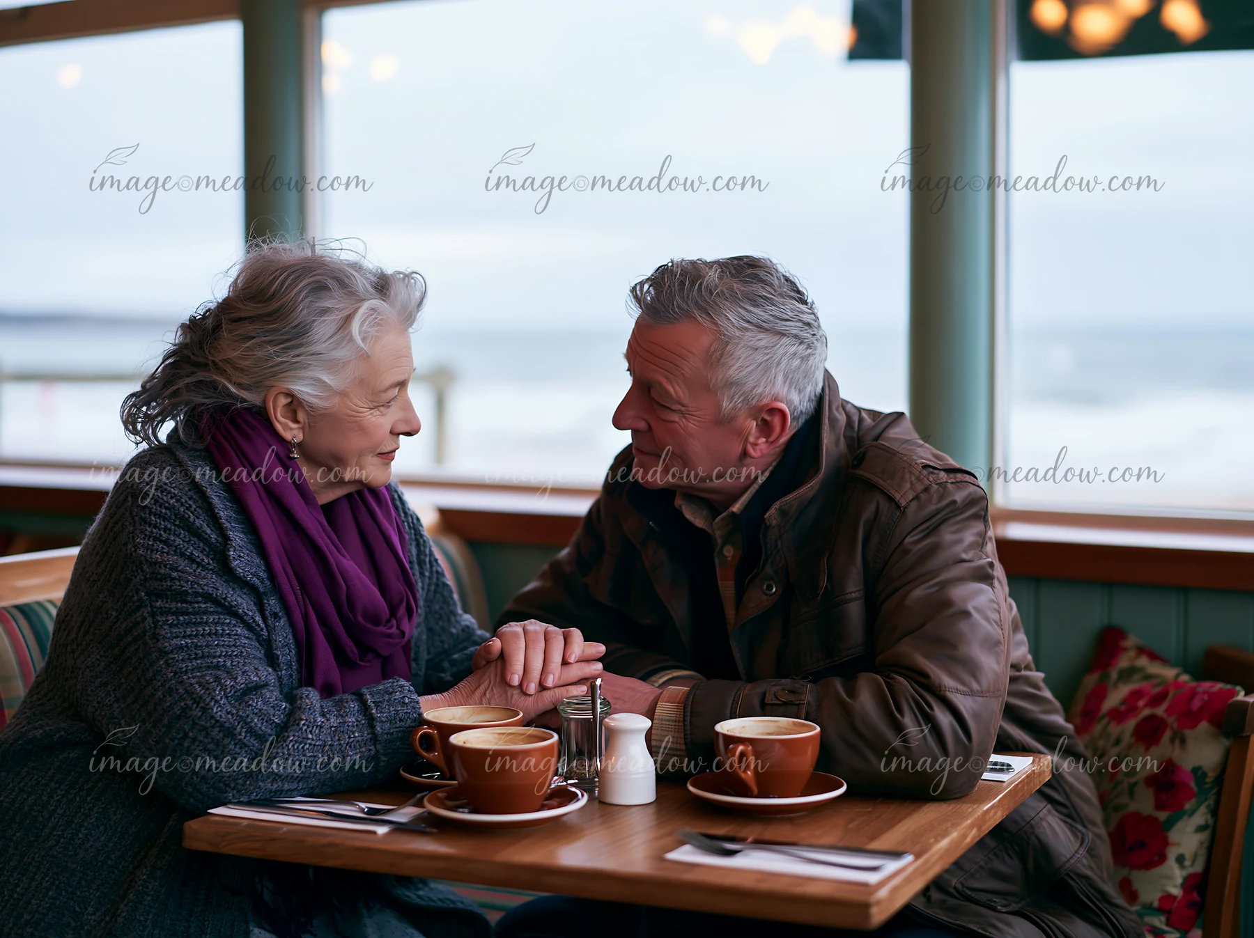 An elderly couple sits together at a table in a cafe, holding hands while gazing into each other’s eyes.