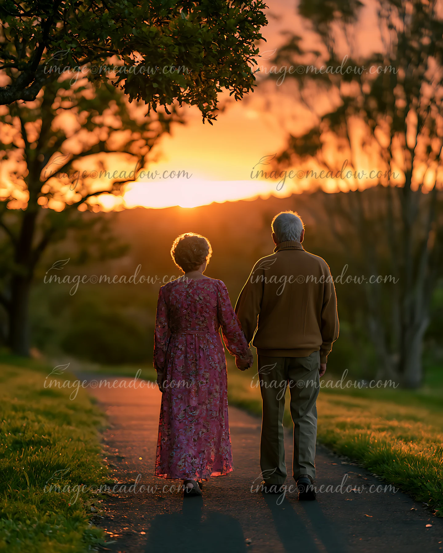 An elderly couple strolls together down a path while holding hands, with a vibrant sunset and trees in the background.