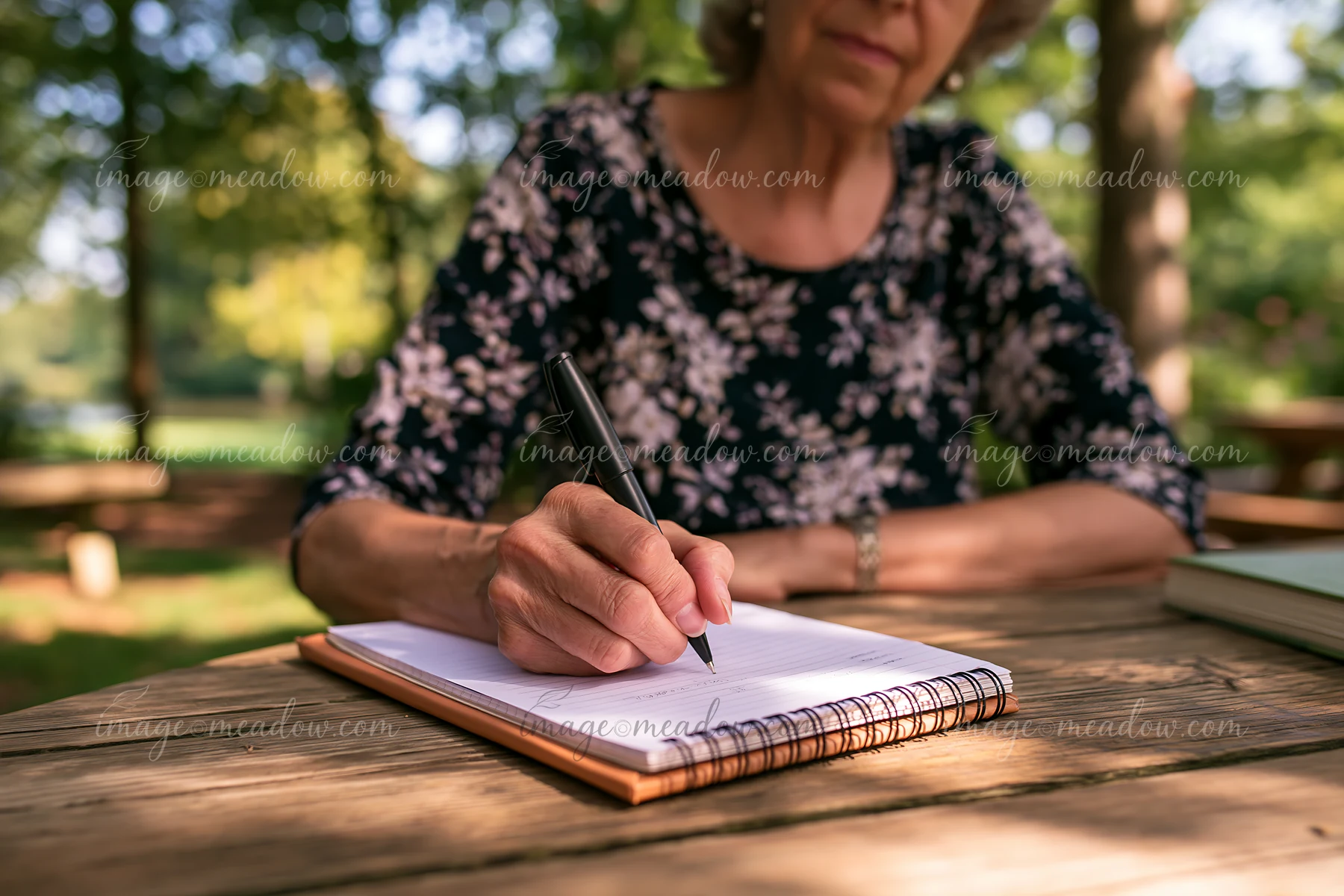 An elderly woman with Alzheimer's writing reminders in a notebook while sitting at a wooden table outdoors.