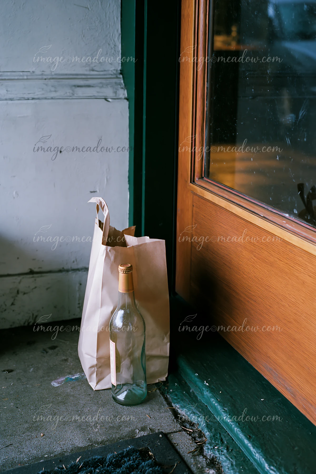 An empty bottle after party sits beside a brown paper bag near a closed wooden door.