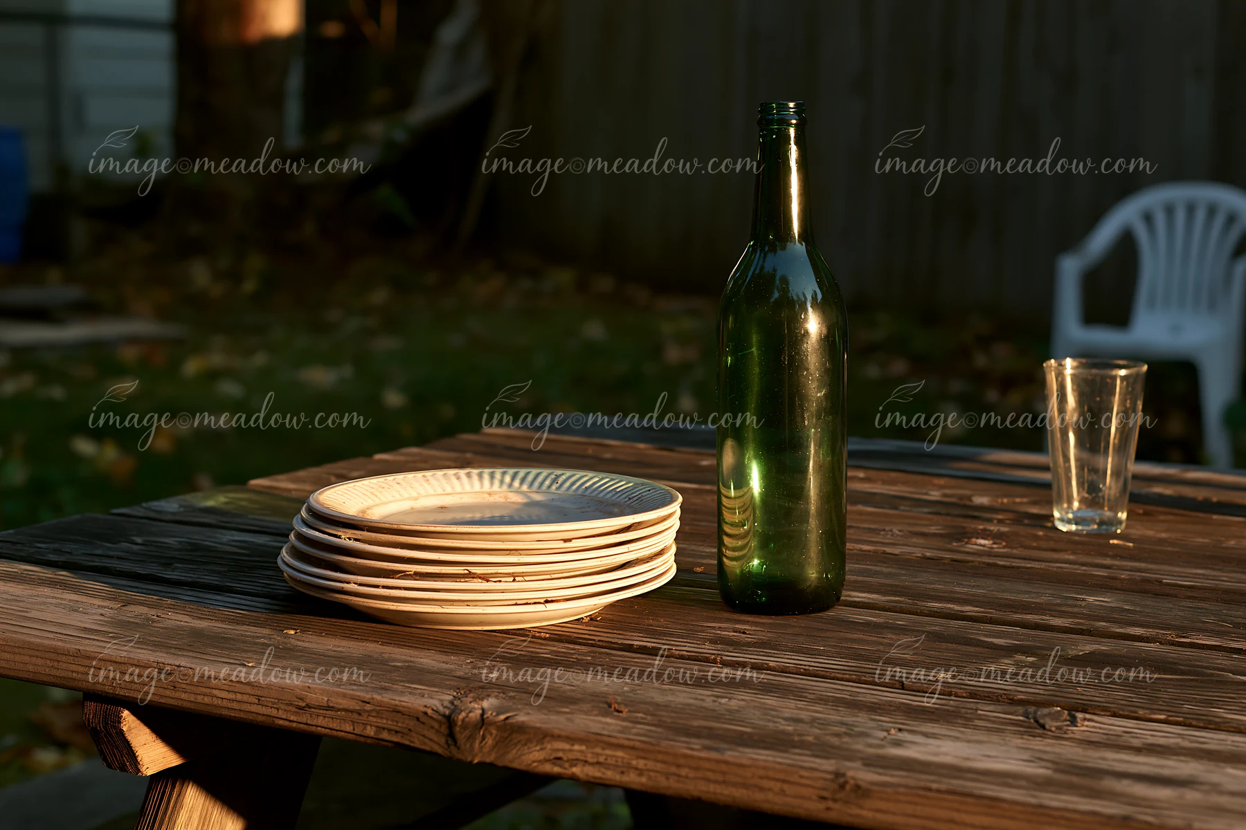 An empty bottle after party stands on a wooden table beside stacked plates and a glass in a backyard setting during evening light.