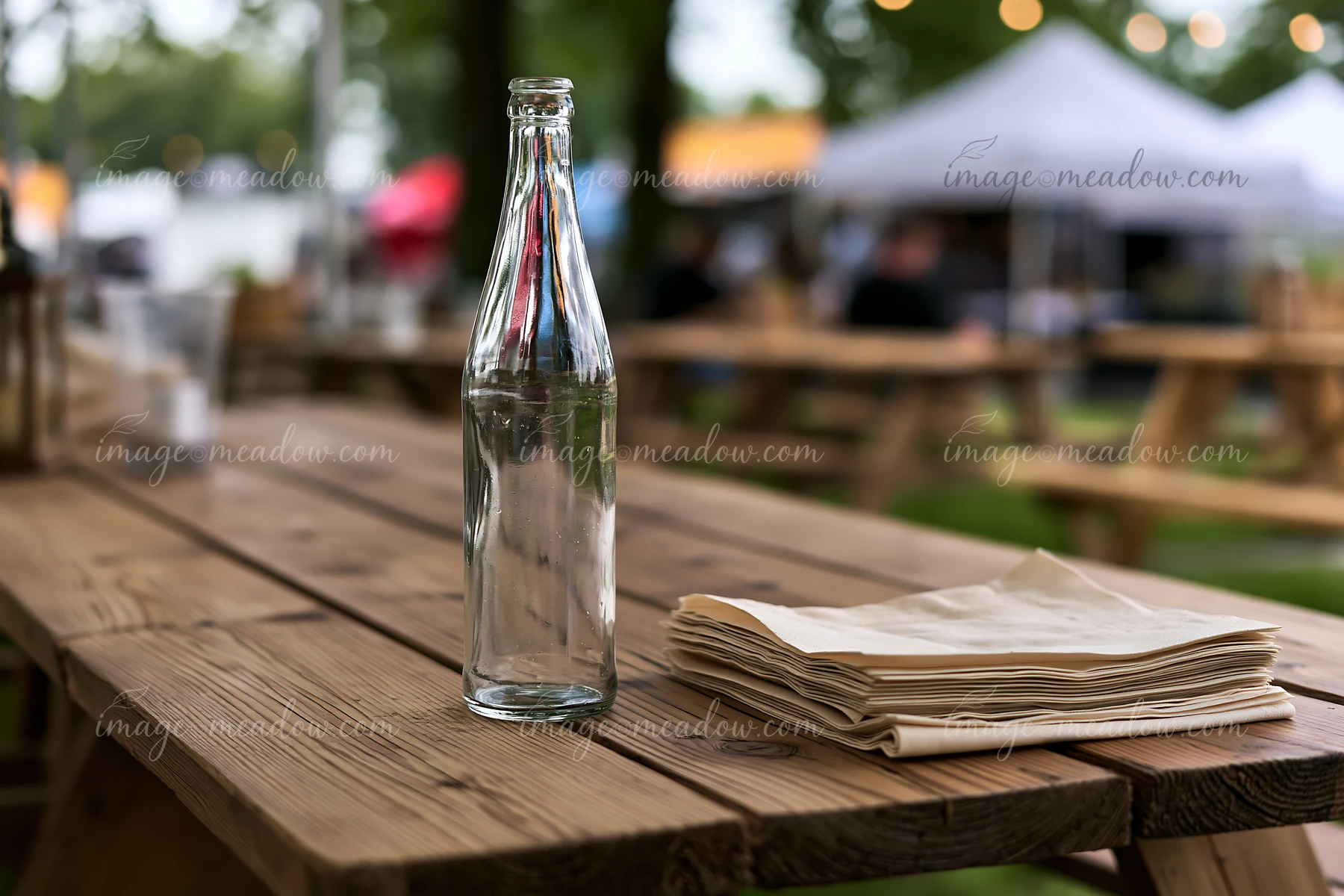 An empty bottle rests on a wooden table next to a stack of napkins during an outdoor event.