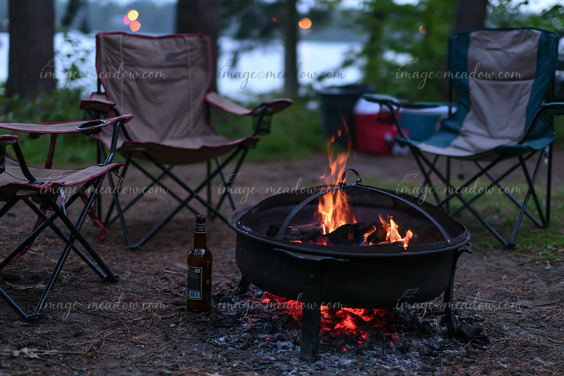 An empty bottle sits beside a campfire in a wooded area.